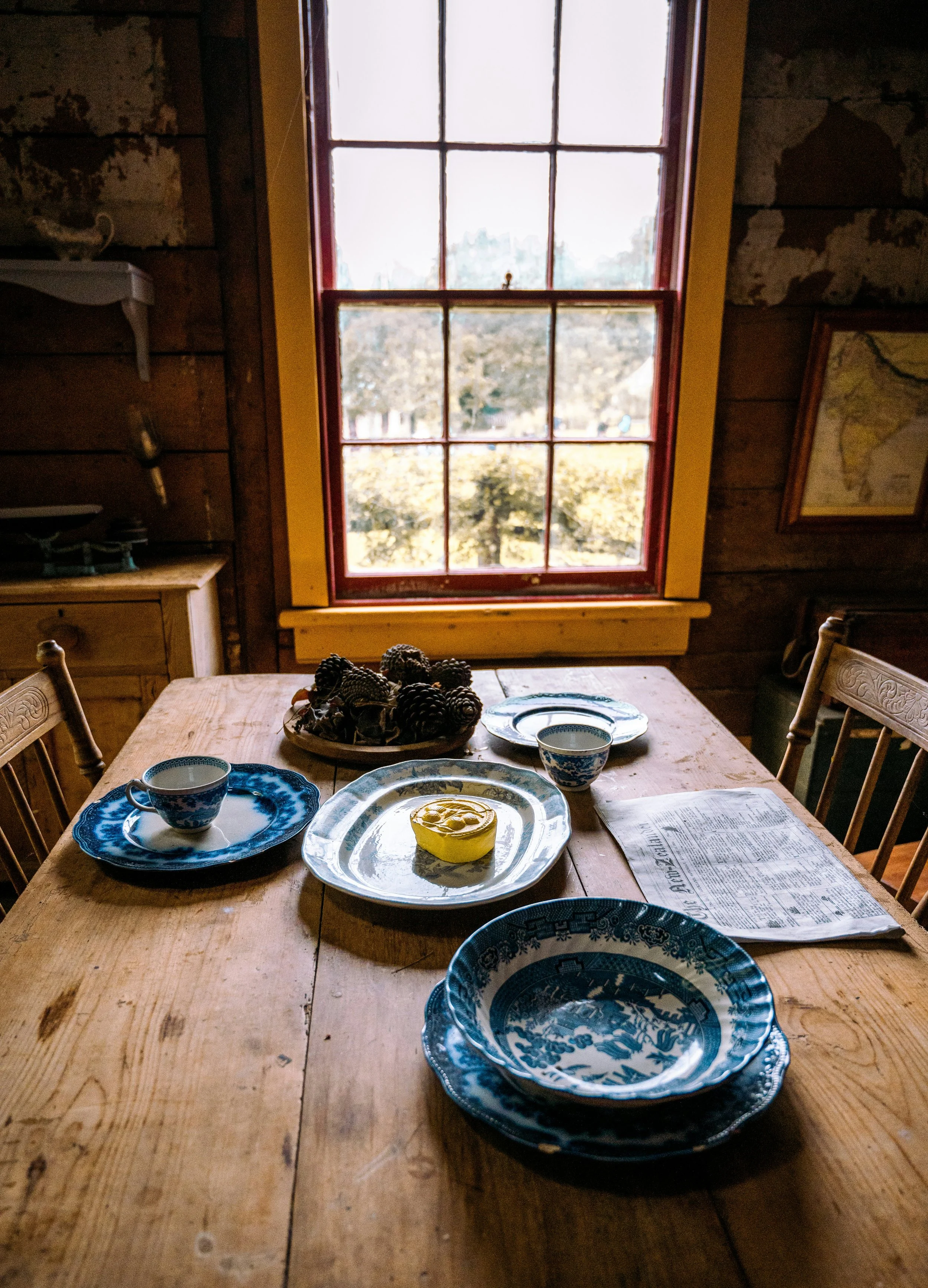 A rustic wooden dining table set with blue and white patterned dishes, a yellow rolled napkin, and pinecones in a bowl, in front of a window with a view of trees outside.