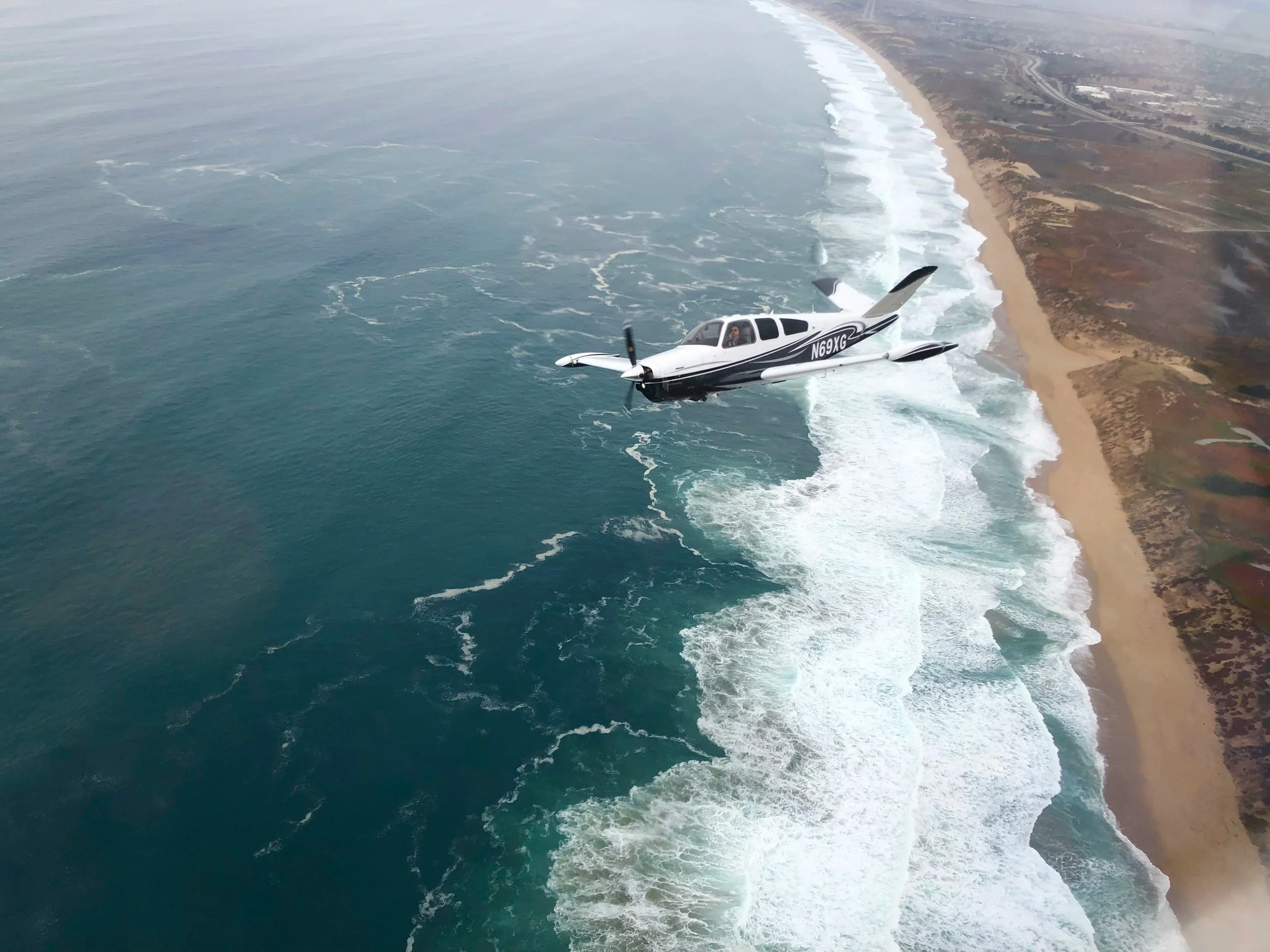 A small white airplane flying over the ocean near a coastline with waves and sandy beach.