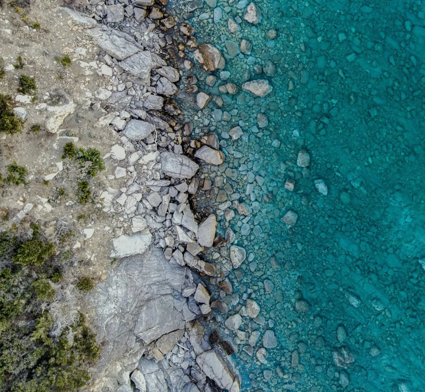 Aerial view of a rocky shoreline next to clear blue water with rocks and some sparse green vegetation.