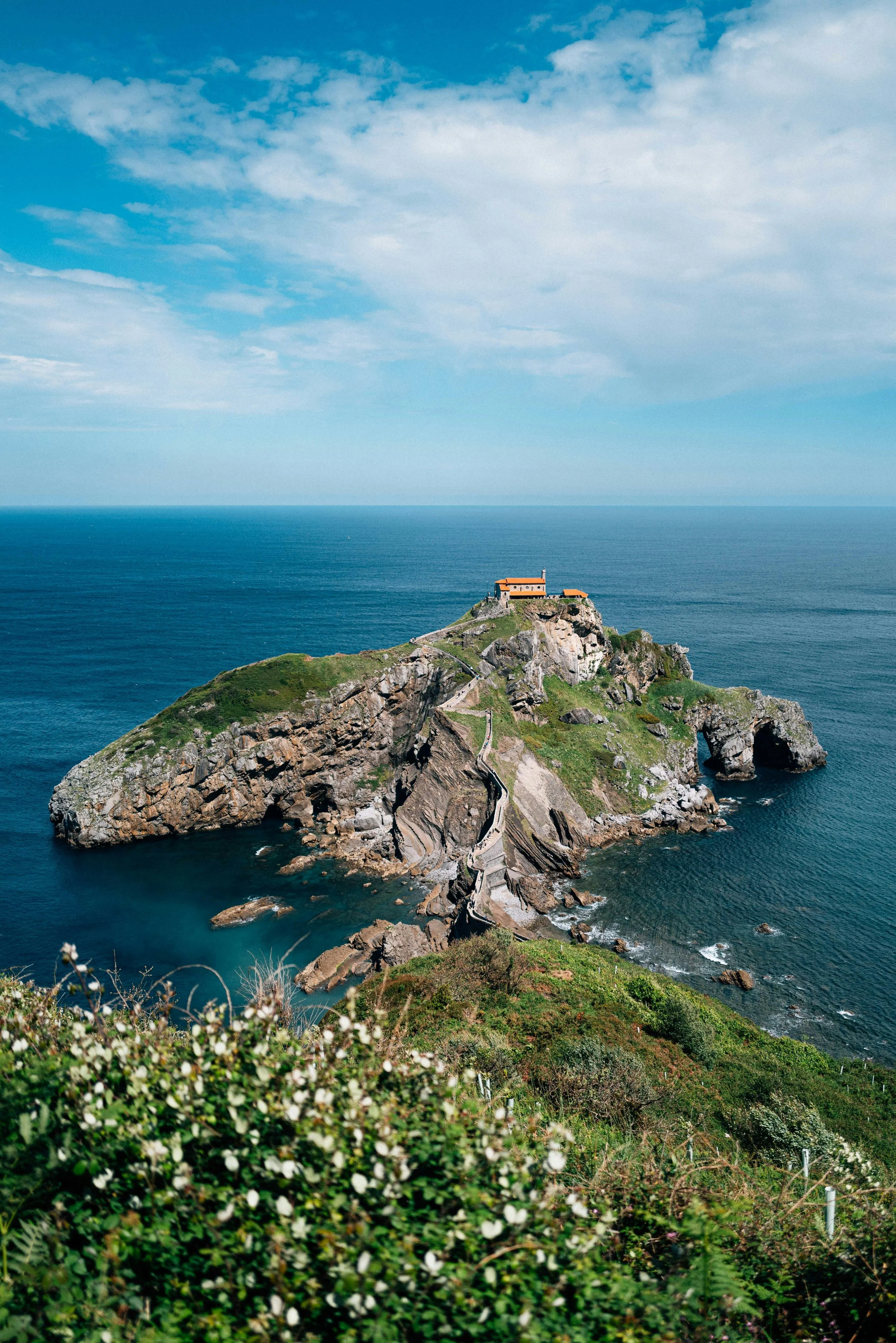 A rocky island with a small building on top, surrounded by the ocean under a partly cloudy sky, with green vegetation and a pathway leading to the structure.
