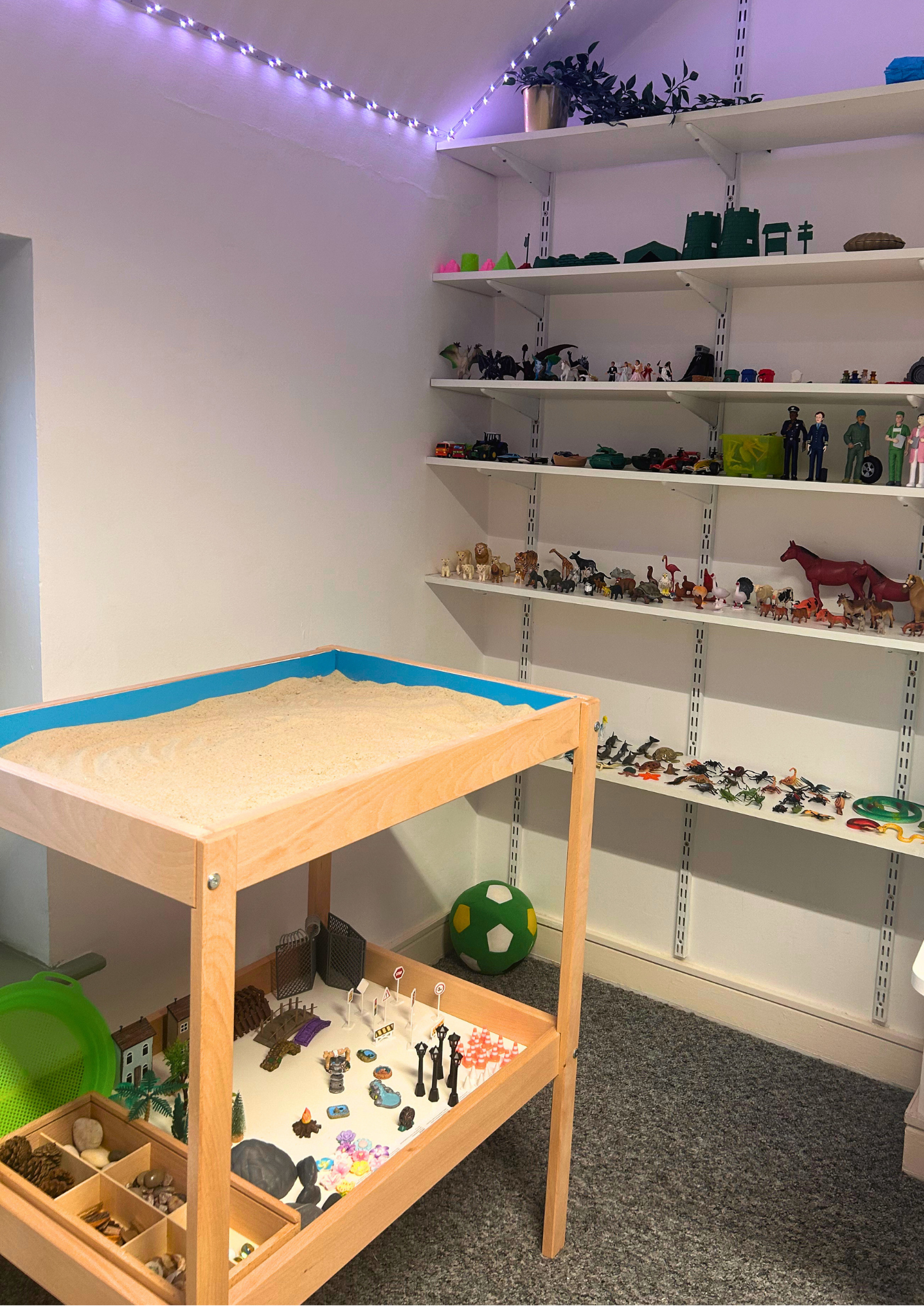 Room with white shelves holding small figurines, toys, and decorative items, a wooden sandbox with sand, a green and white ball, and a display table with small collectible items, in a space lit with purple string lights.