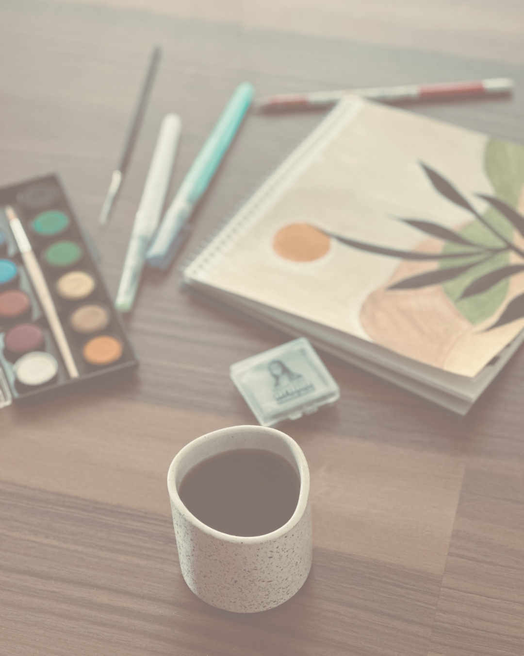 A table with watercolor painting supplies, including paint set, brushes, a sketchbook, and a cup of coffee.