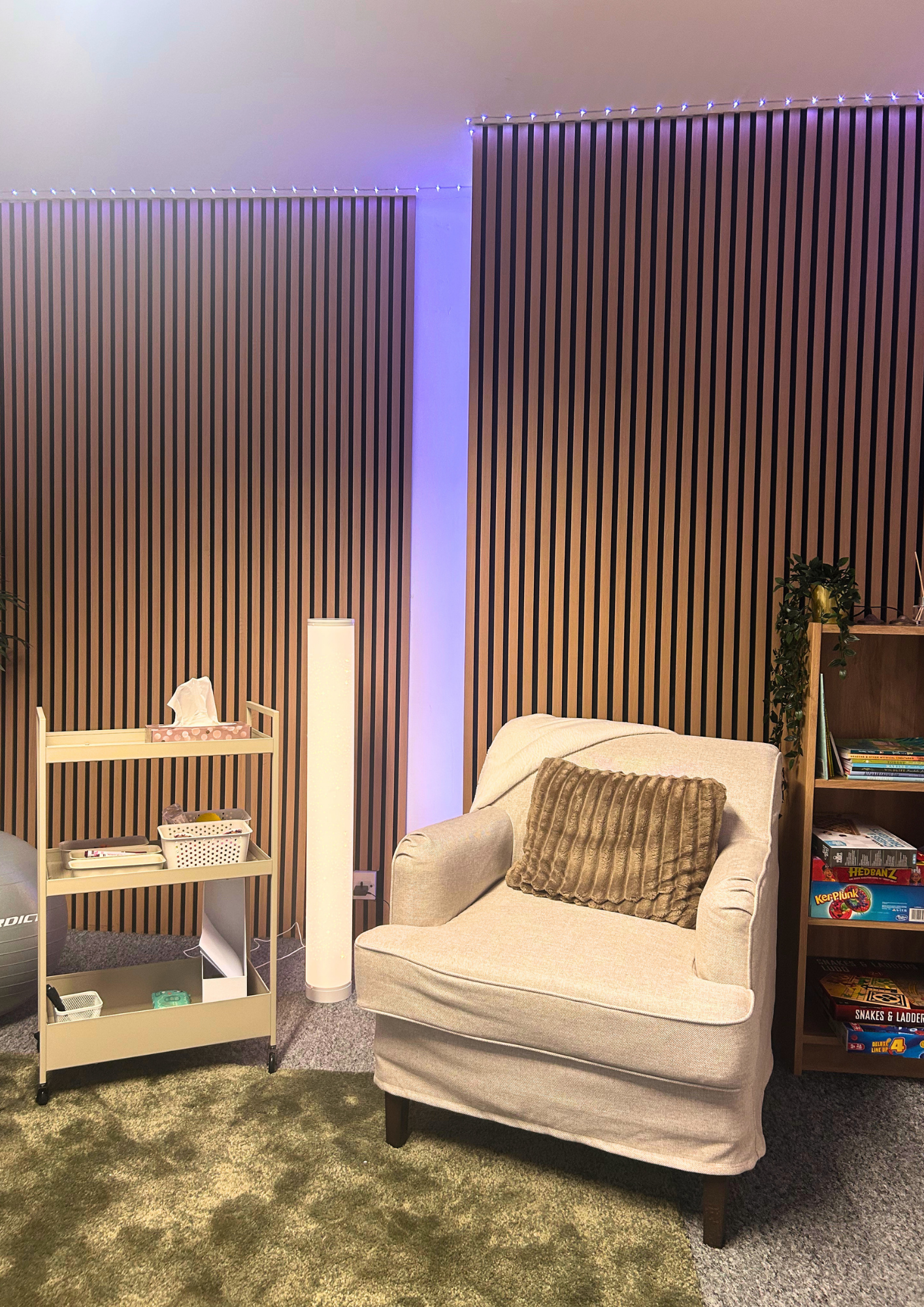 Living room corner with a beige armchair, a brown textured pillow, a wooden bookshelf with board games, a white rolling cart with containers, a tall white lamp, and decor elements on a green and gray carpet. Brown wooden slat wall with blue string lights in the background.