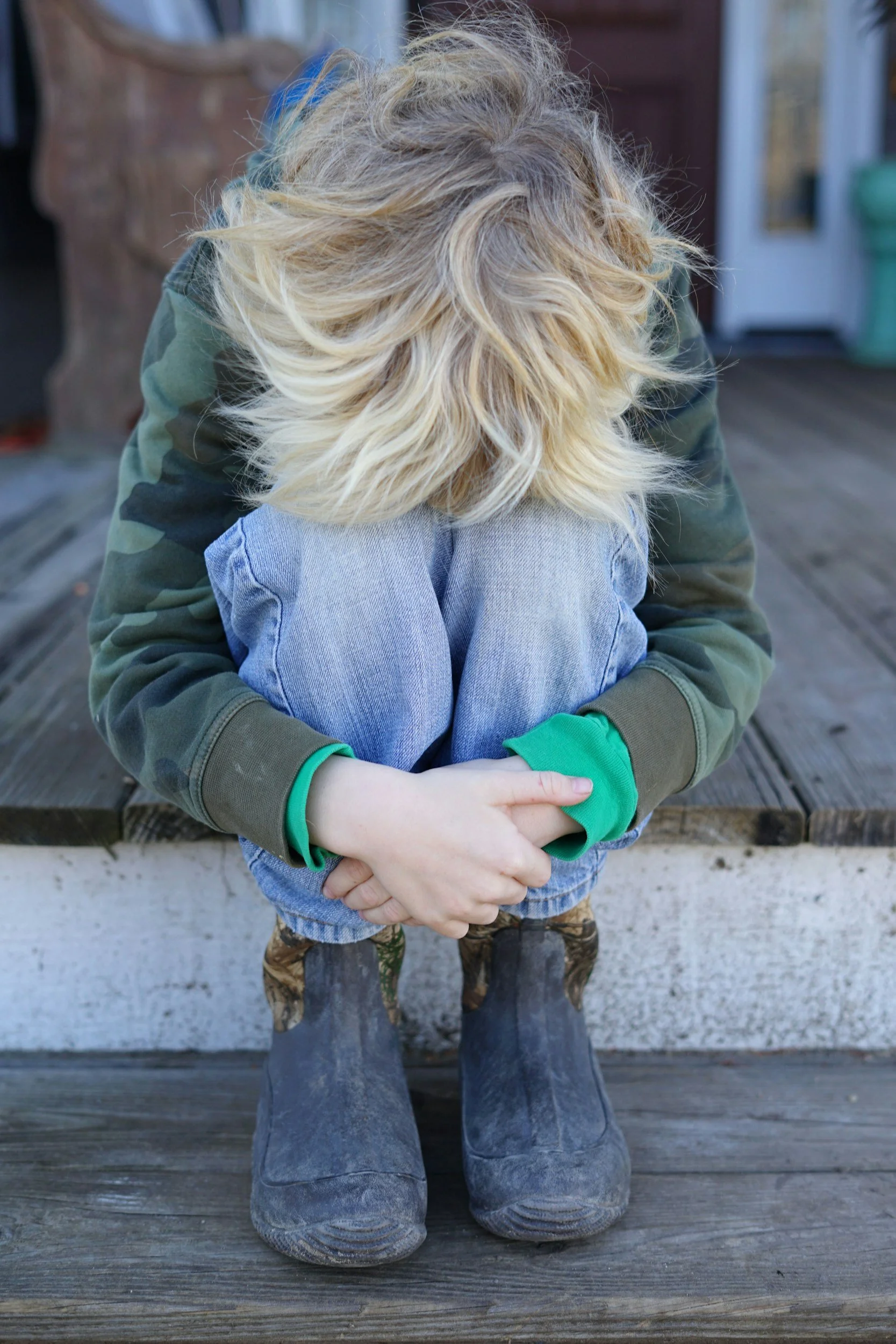 A person with blonde, tousled hair, wearing a green camouflage jacket and blue jeans, sitting on a wooden bench with their head down, arms crossed and hands clasped, on a wooden deck with a blurred background.