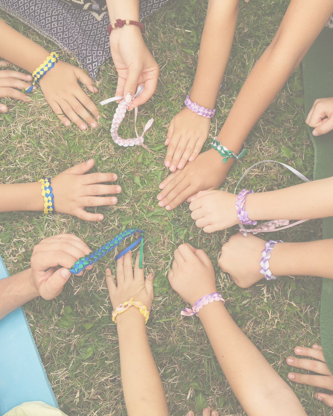 Multiple children with colorful braided friendship bracelets on their wrists, reaching towards the center on grass.