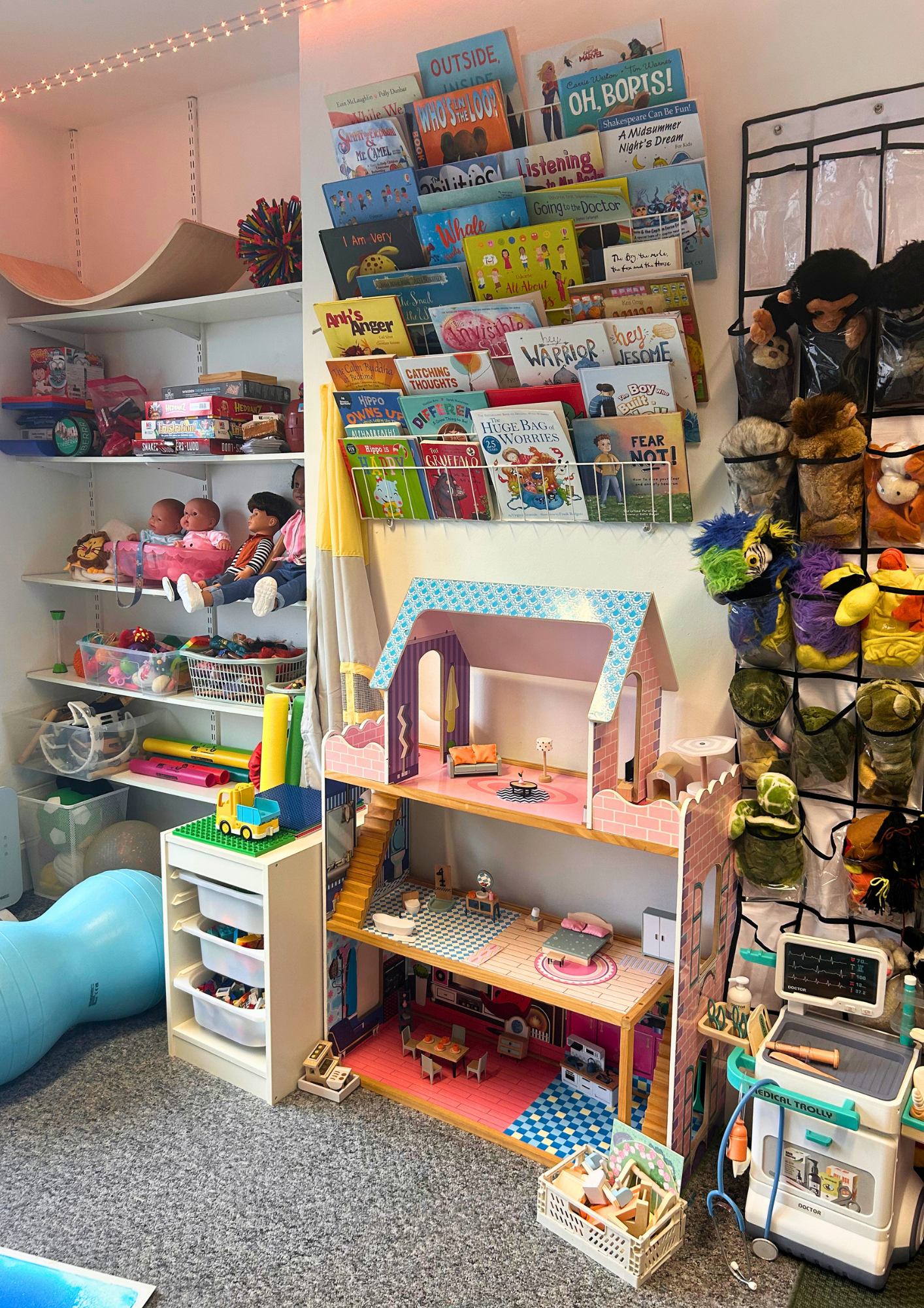 A playroom with toy shelves filled with dolls, stuffed animals, books, and a dollhouse. There are toys on the floor and a medical toy cart on the right.