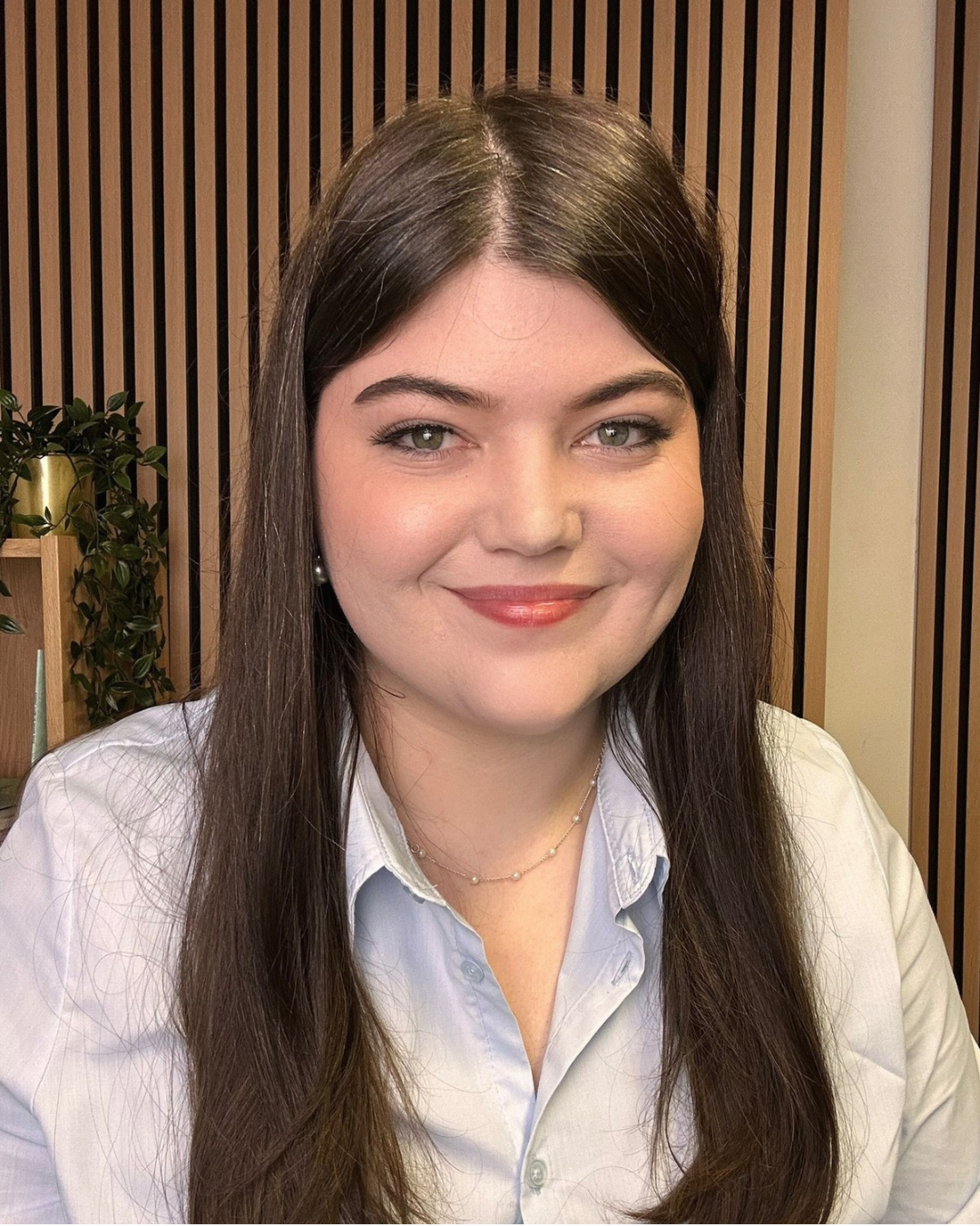 A woman with long brown hair and light green eyes, smiling, wearing a white button-up shirt, pearl earrings, and a delicate chain necklace, in front of a wooden slat wall and a potted plant.