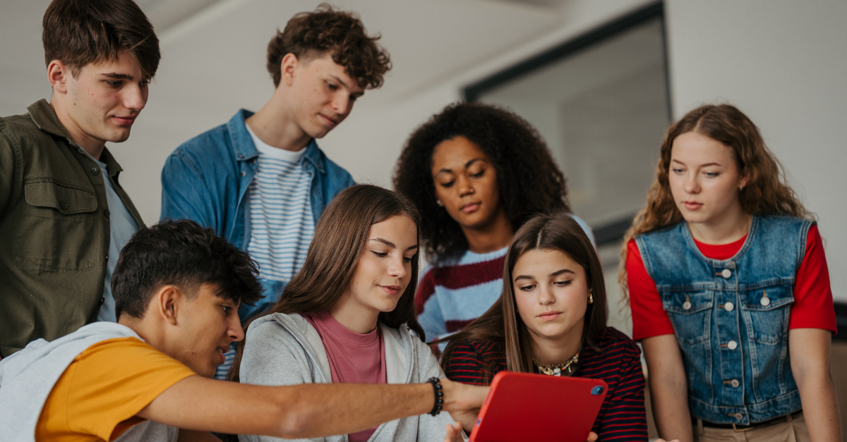 A group of seven teenagers gathered around a tablet, looking at the screen together in a classroom or study area.