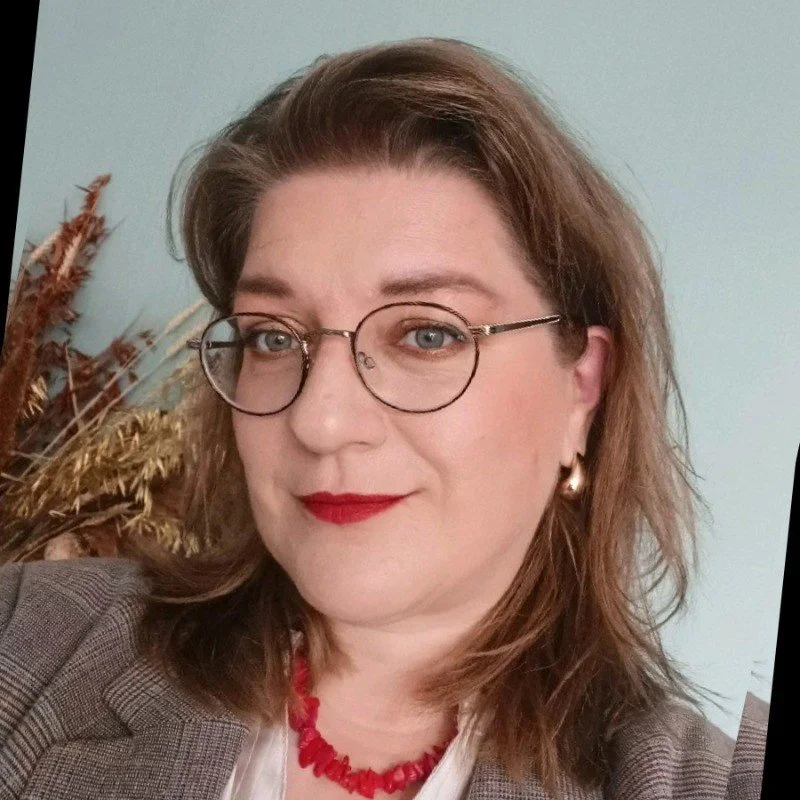 A woman with shoulder-length wavy brown hair, wearing glasses, red lipstick, a red necklace, earrings, and a gray checkered blazer, posed indoors with dried plants in the background.