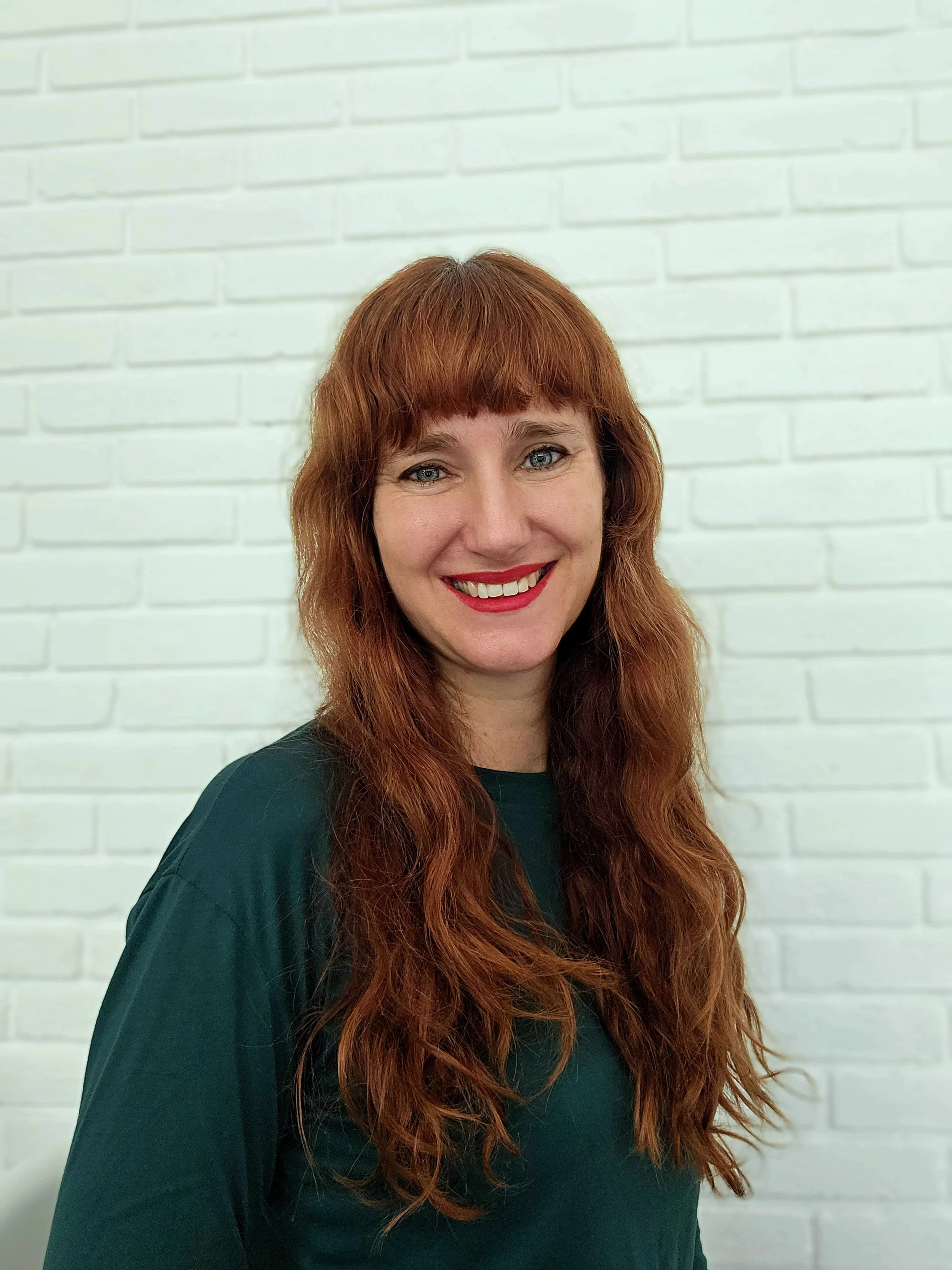 A woman with long red hair and blue eyes smiling, wearing a dark green shirt, standing in front of a white brick wall.