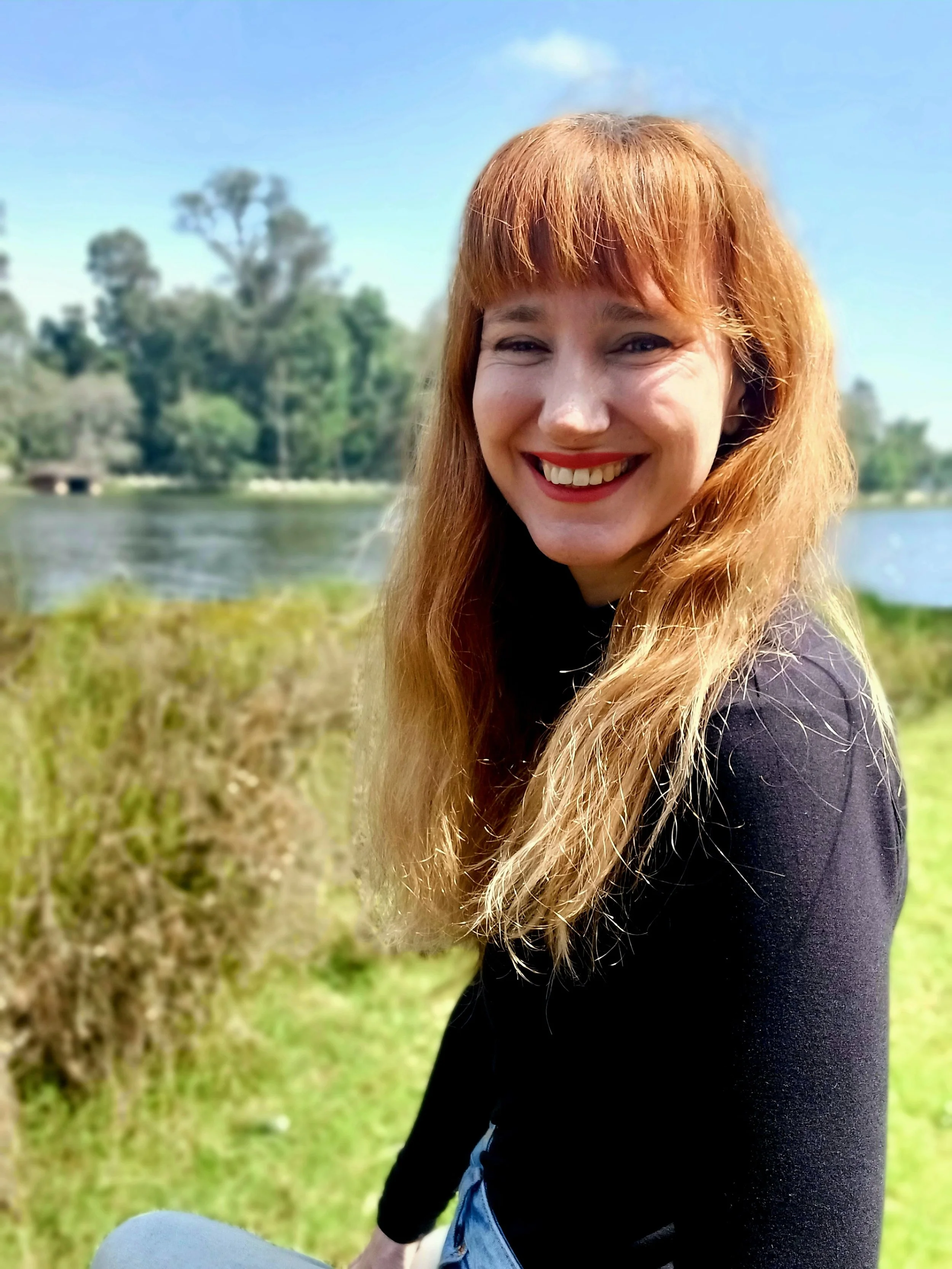 A woman with long red hair, wearing a black top, smiling outdoors near a river, with trees and clear blue sky in the background.