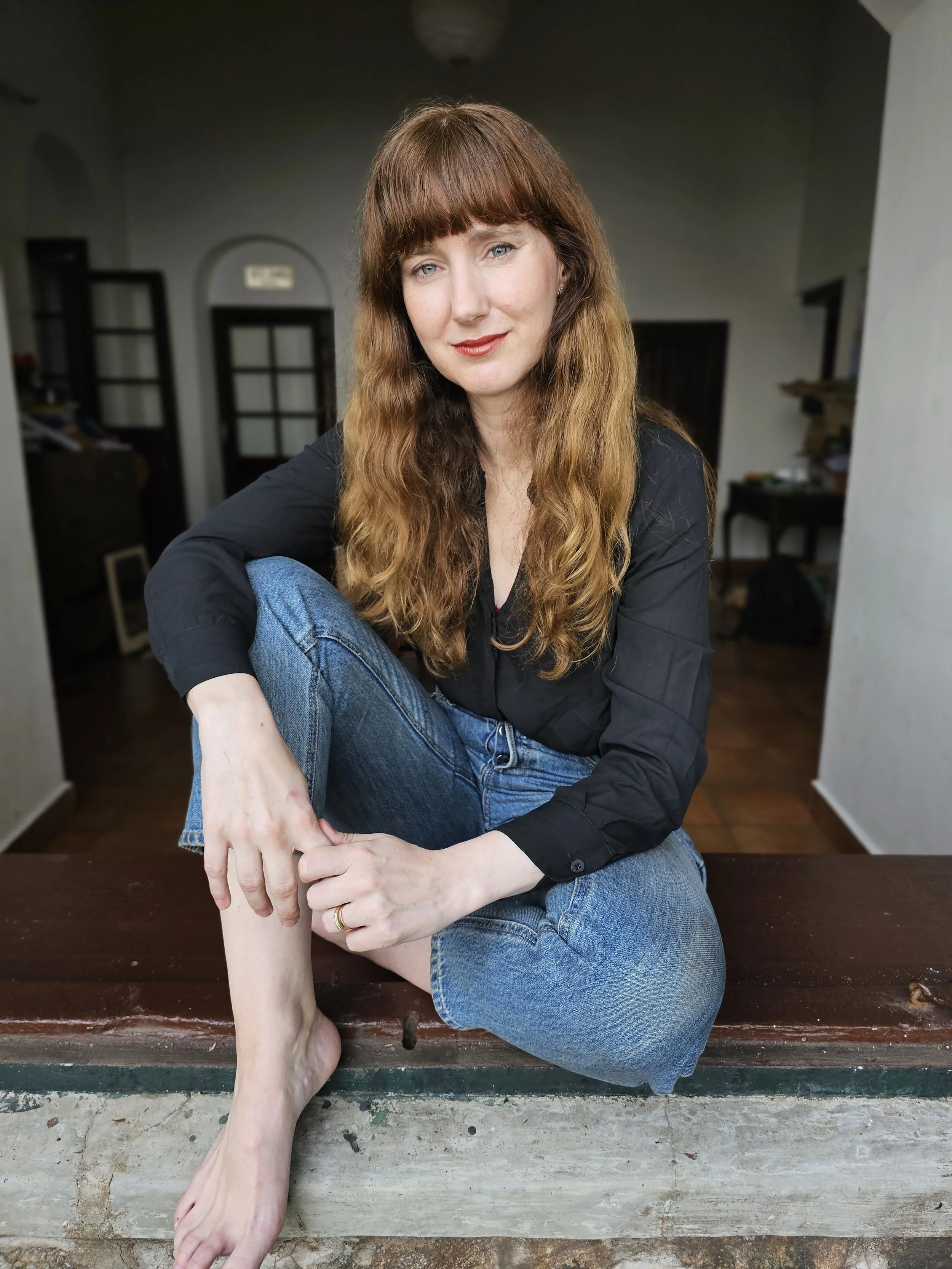 Woman with long reddish hair, wearing a black shirt and jeans, sitting on a wooden bench inside a room, with a person’s foot touching her arm in the foreground.