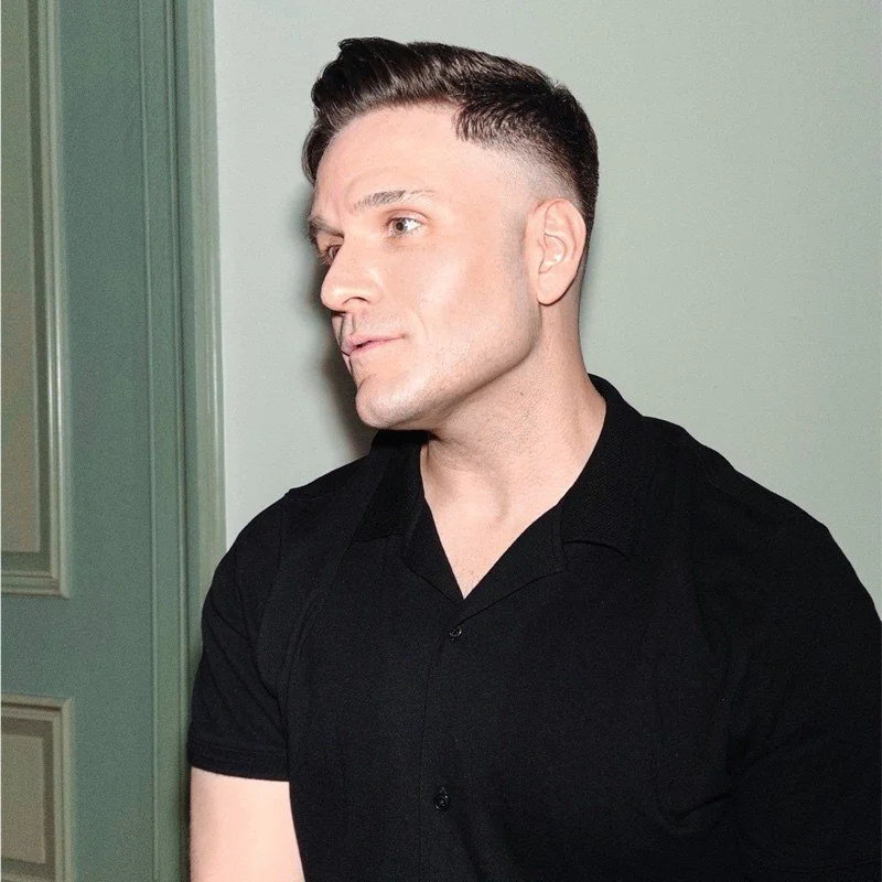 A man with a modern undercut hairstyle wearing a black shirt, standing indoors against a plain wall.