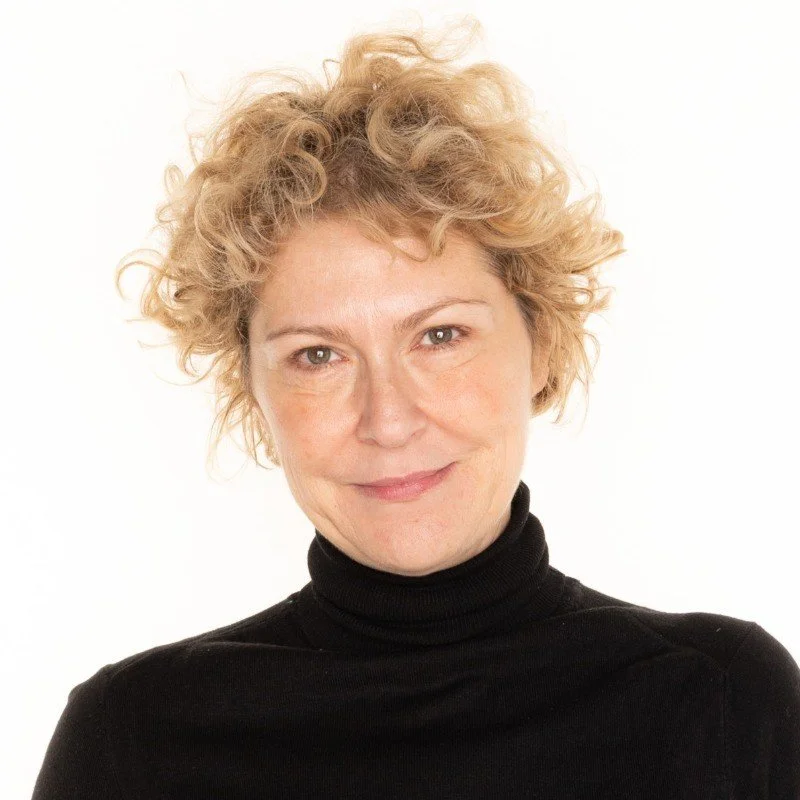 A portrait of a woman with curly blonde hair, wearing a black turtleneck, smiling subtly against a plain white background.