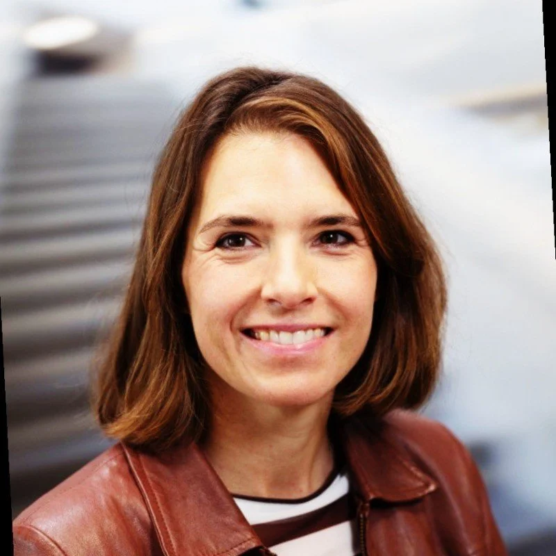 Close-up of a smiling woman with shoulder-length brown hair wearing a brown leather jacket and striped shirt, outdoors with blurred background.