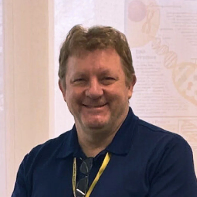 A smiling middle-aged man with light brown hair and a beard, wearing a navy blue shirt and glasses hanging from a yellow lanyard around his neck, standing in front of a light-colored wall with faint text and images.