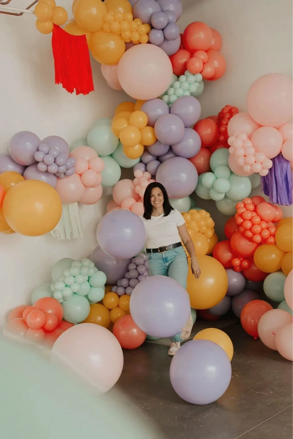 A woman smiling and posing among colorful balloon decorations, with balloons in pastel shades of purple, pink, yellow, green, and orange covering a wall.