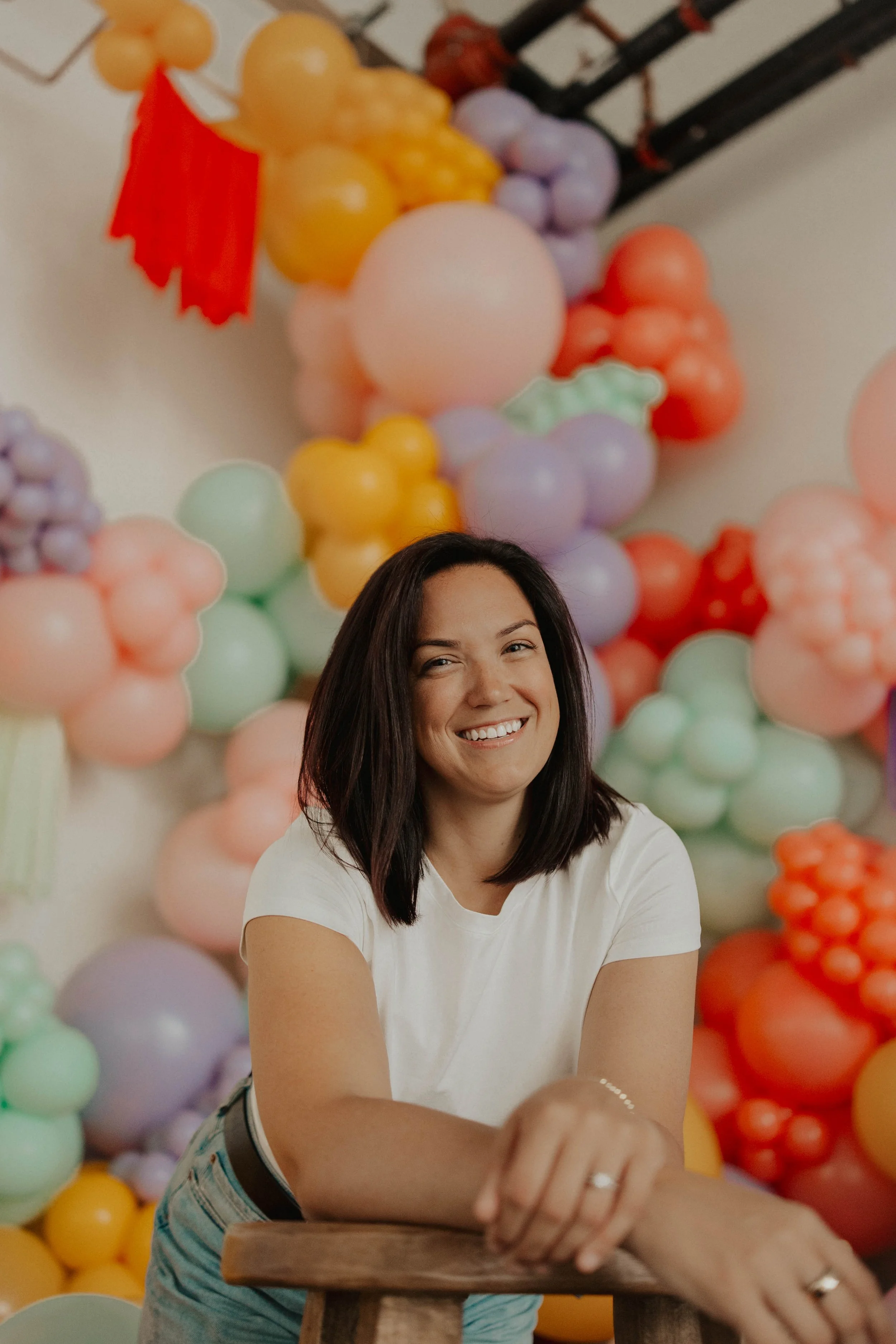 A smiling woman with shoulder-length dark hair, wearing a white t-shirt, sitting at a table in front of a colorful balloon wall. The balloons are in various pastel colors and arranged to resemble a fruit or garden scene. She is looking at the camera, with her left arm resting on the table.
