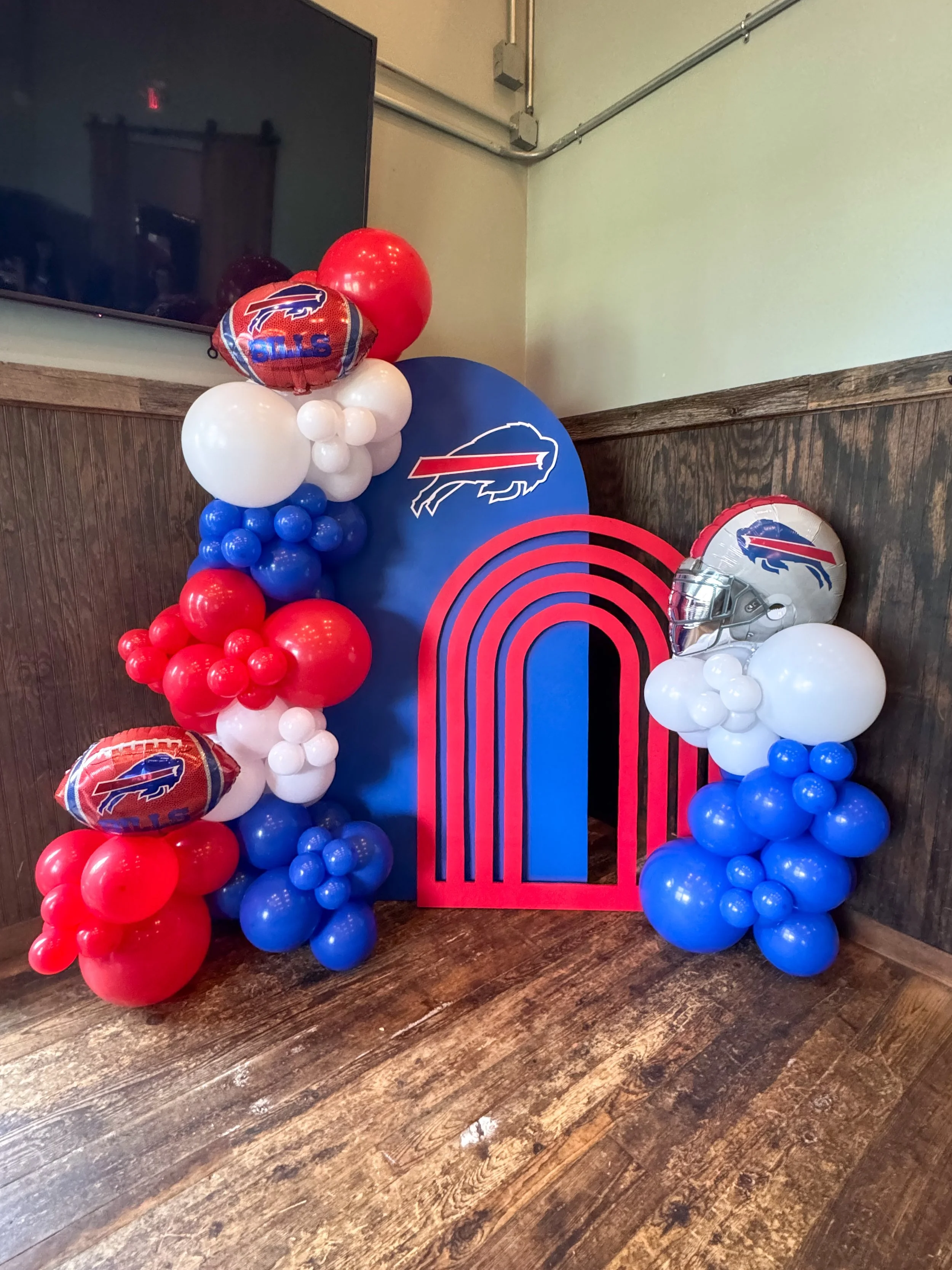Decorative display featuring an arch of red, white, and blue balloons, with a large blue cutout of a buffalo logo and target design, and two balloons shaped like footballs with the Buffalo Bills logo in an indoor setting.