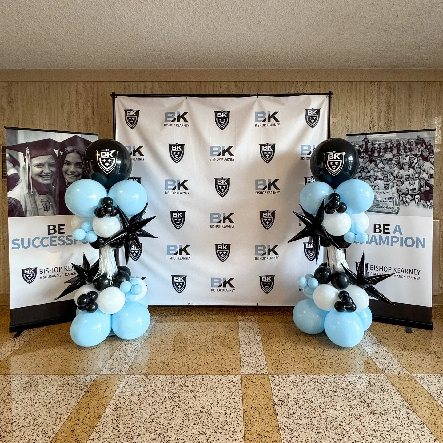 Wedding or graduation celebration setup with black, light blue, and white balloons, decorated with black star-shaped balloons, in front of a backdrop with the Bishop Kearney school logo. Two vertical banners feature black and white photos with motivational messages for students.