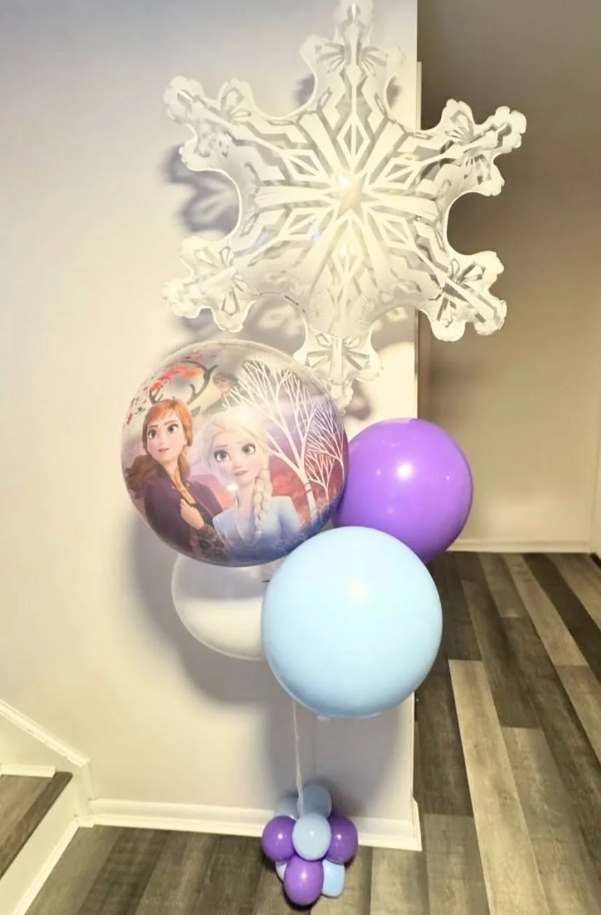 A balloon arrangement with a large snowflake-shaped decoration, a 'Frozen' themed balloon featuring Anna and Elsa, and several purple and light blue balloons anchored with smaller purple and blue balloons at the base.