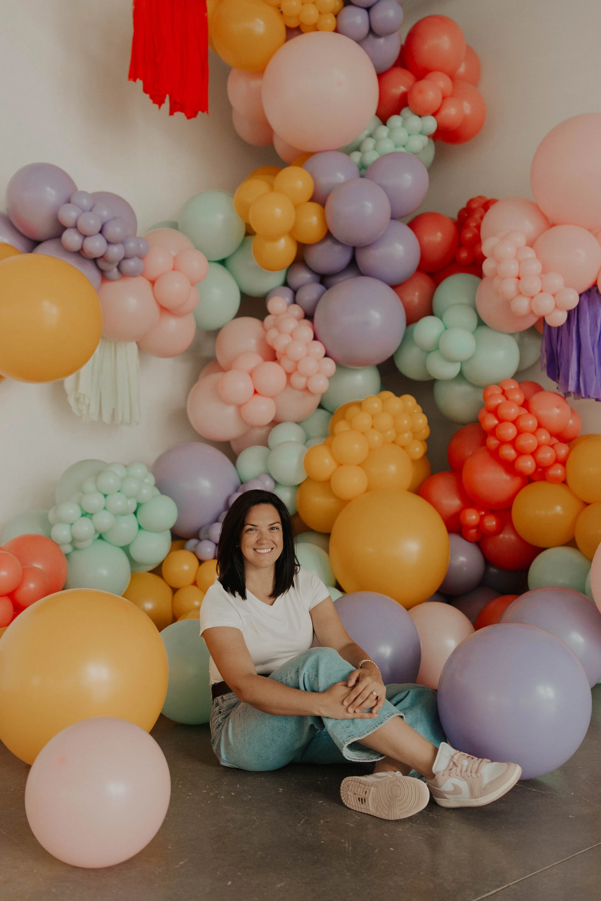 A woman sitting on the floor in front of a colorful balloon arrangement, smiling at the camera.