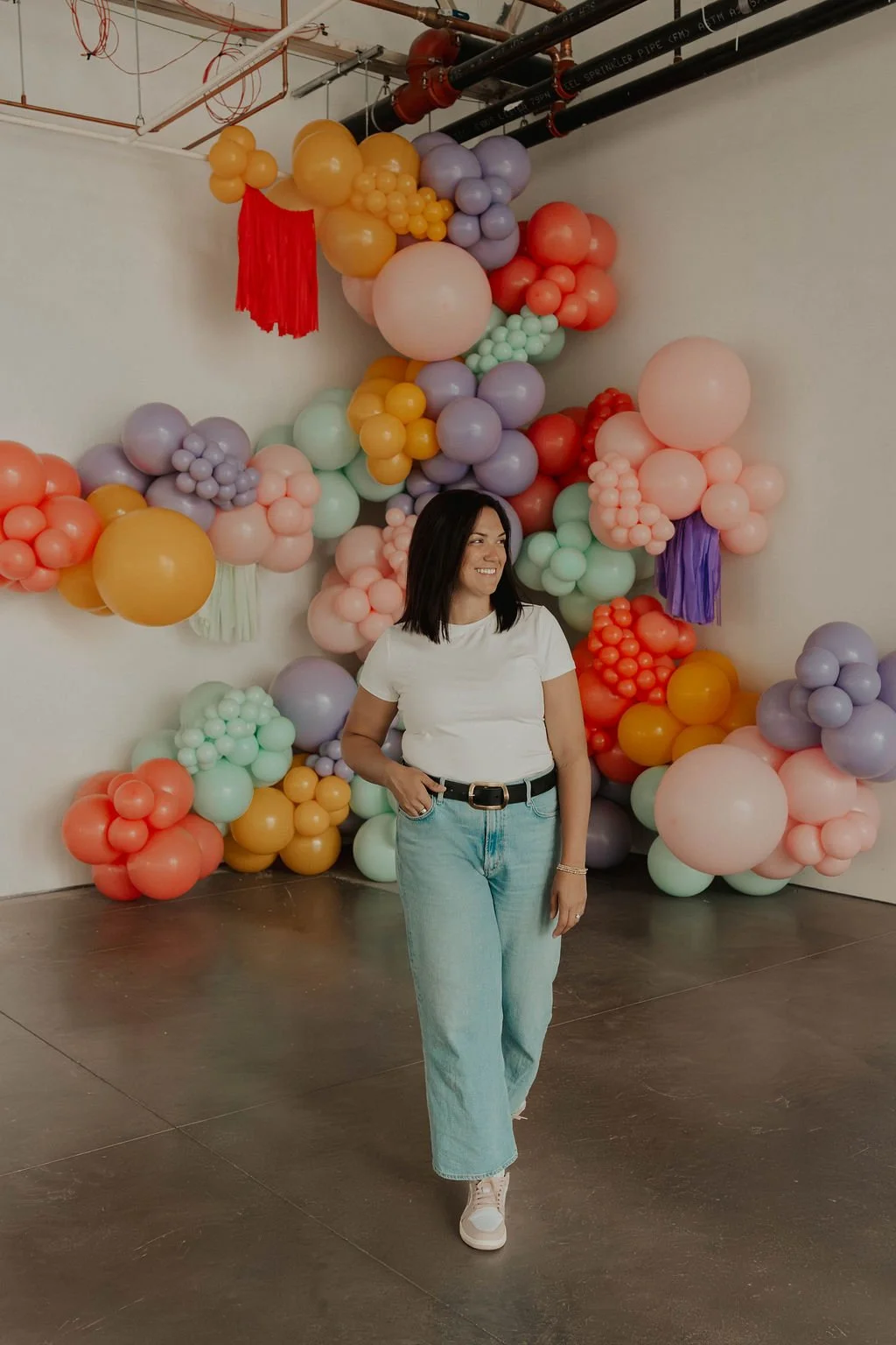 A woman with shoulder-length dark hair wearing a white t-shirt and light blue jeans smiling while standing in front of a colorful balloon installation with various pastel and bright balloons arranged in clusters on a white wall.