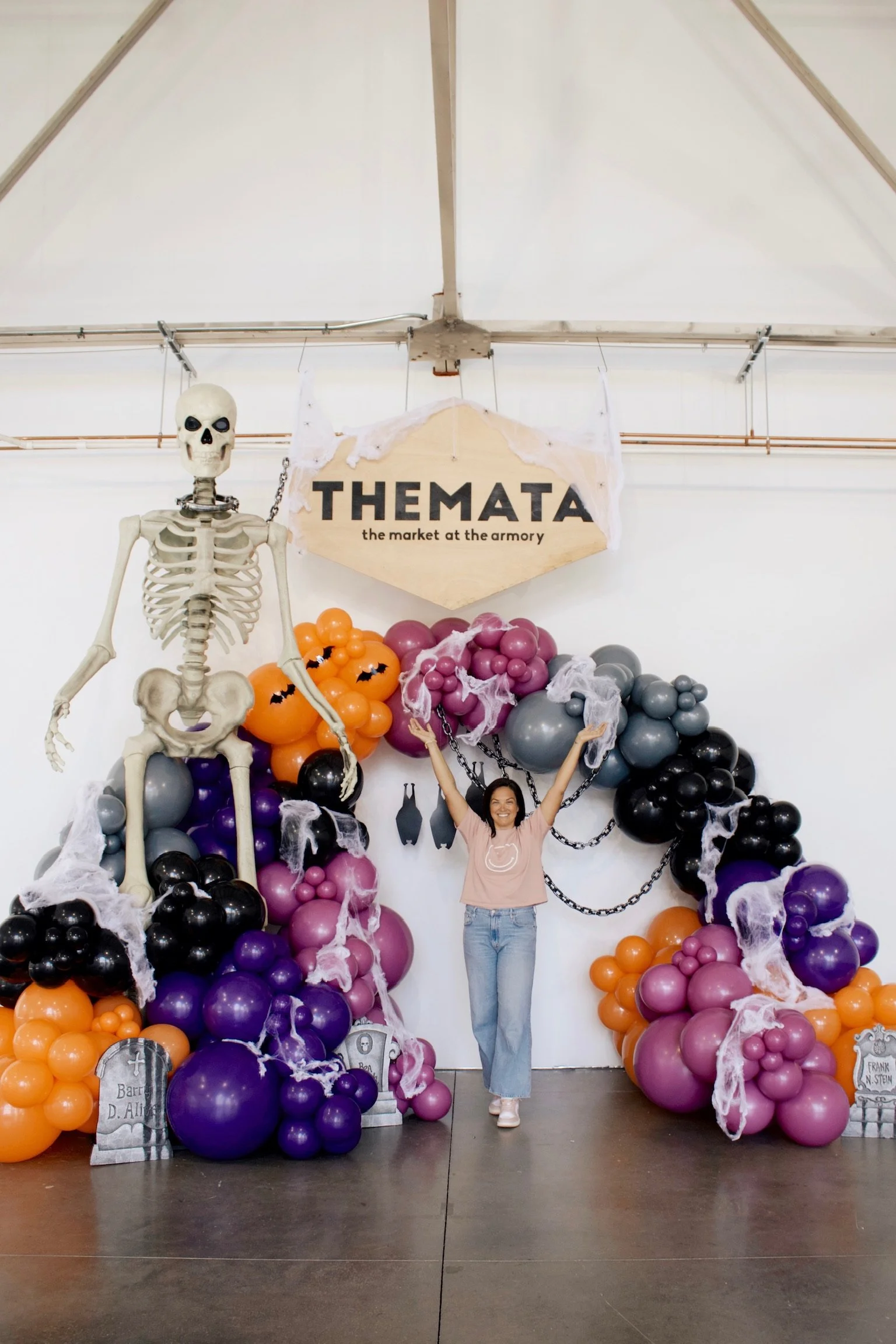 A woman smiling and standing with arms raised in front of a Halloween themed decoration with skeleton, orange, purple, and black balloons, fake spider webs, and tombstone props, with a sign that reads 'THE MATA, the market at the armory'.