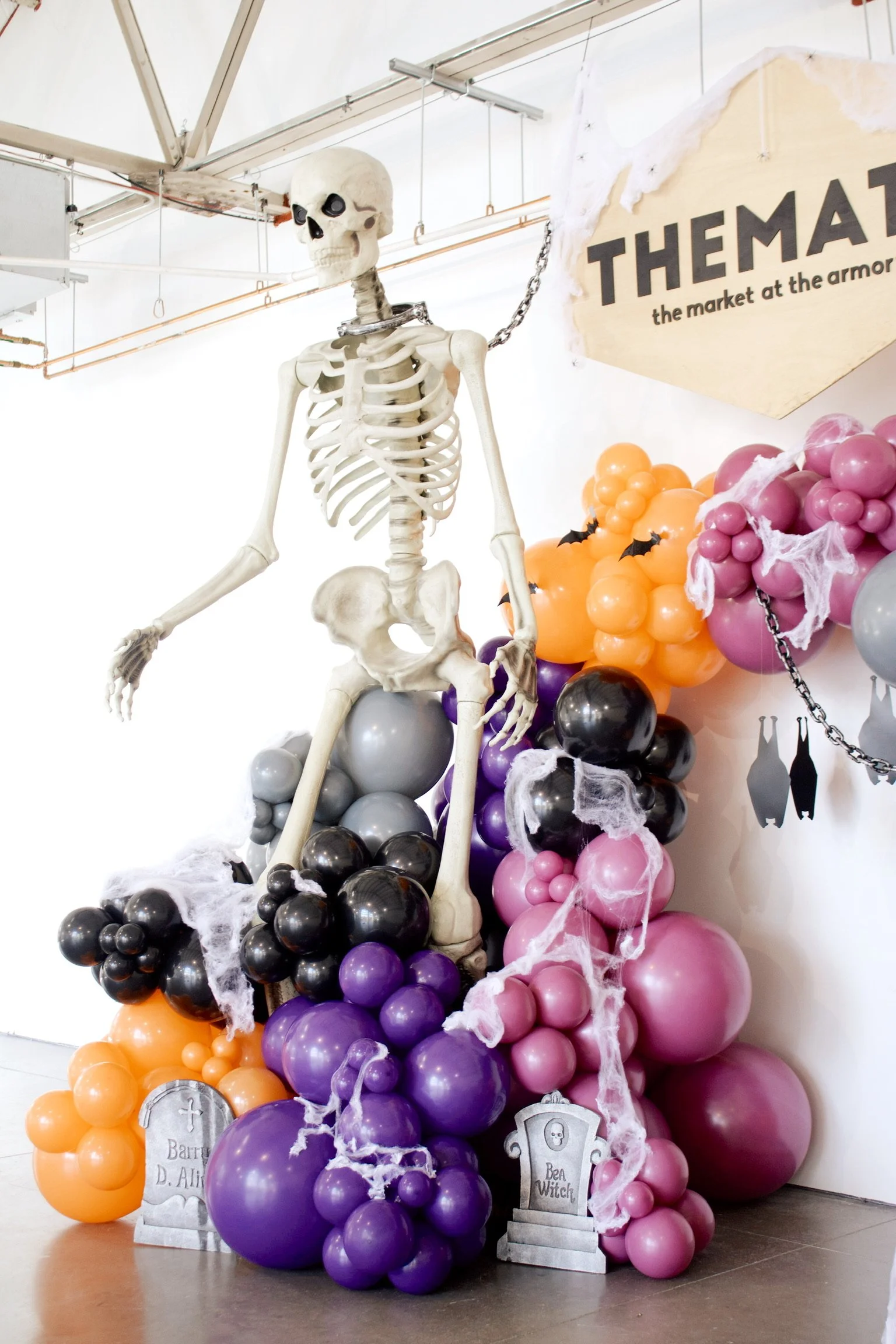 A Halloween-themed display featuring a life-size skeleton decoration surrounded by purple, pink, orange, and black balloons, some with fake cobwebs, and graveyard signs with names like 'Ben Witch' and 'Barrett D. Alice'.