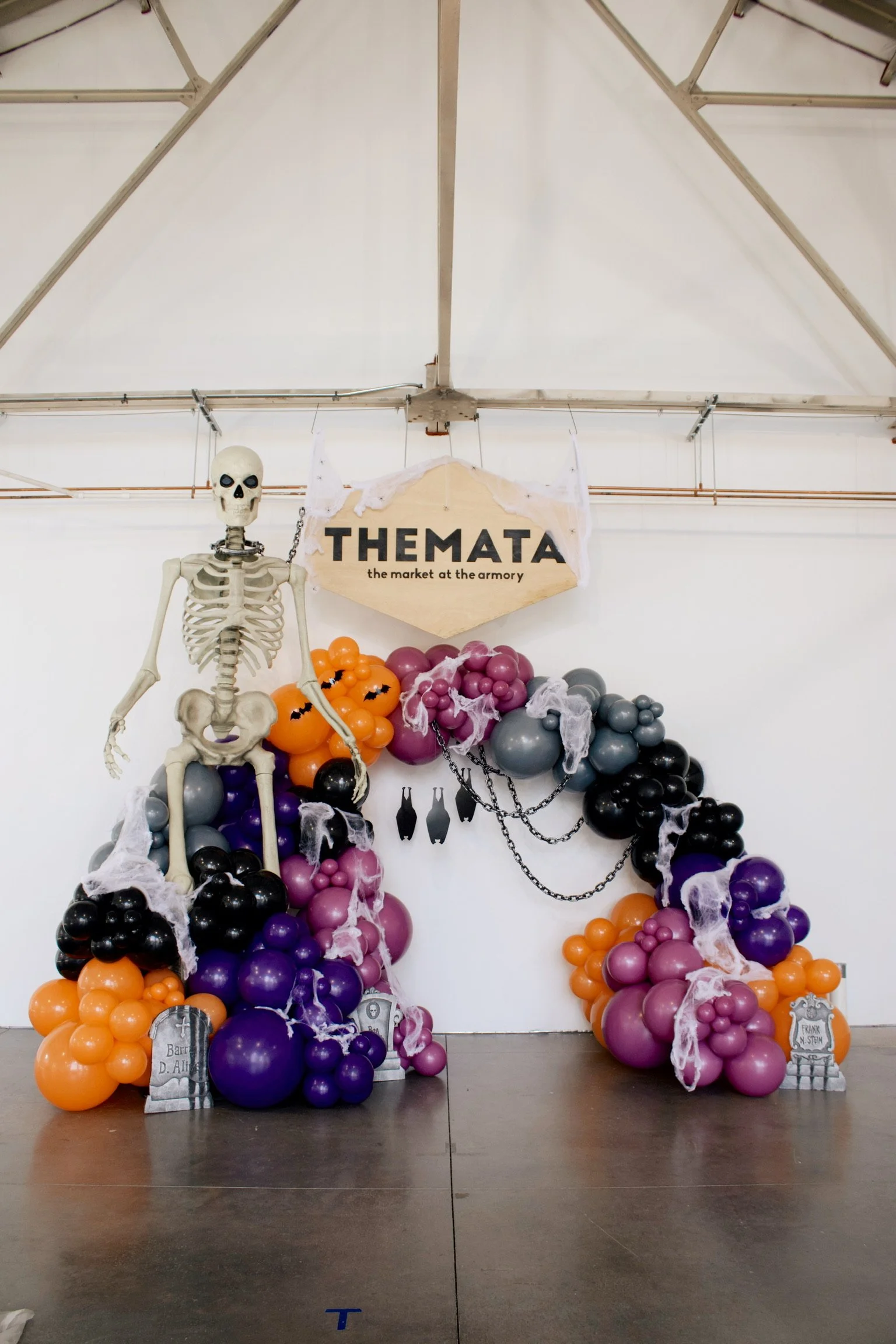 Halloween-themed display with a smiling skeleton hanging among orange, purple, black, and gray balloons, and a sign that reads 'THE MATA the market at the armory'.