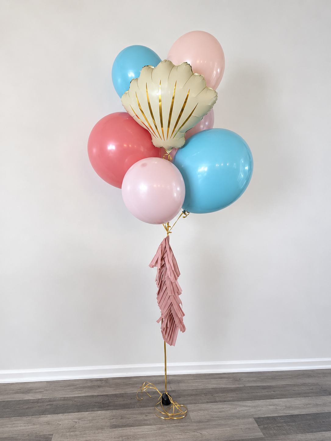 Cluster of pink, blue, and white balloons with a gold and cream shell-shaped foil balloon, tied to pink paper fringe streamer on a light hardwood floor and plain white wall.