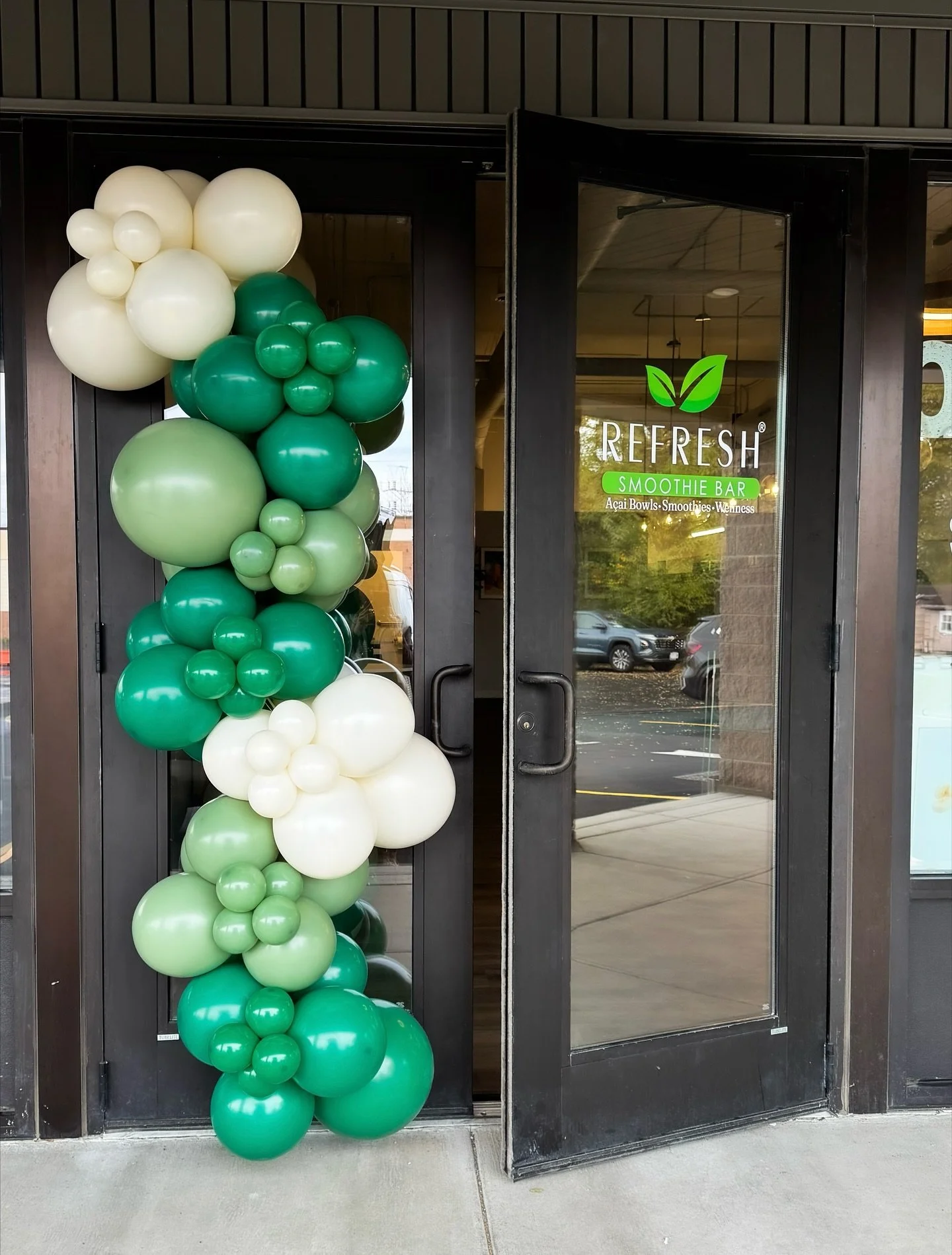 A door decorated with green and white balloons arranged in a cluster on the left side. The door has a sign that reads 'REFRESH Smoothie Bar' with a leaf logo, indicating a smoothie shop.