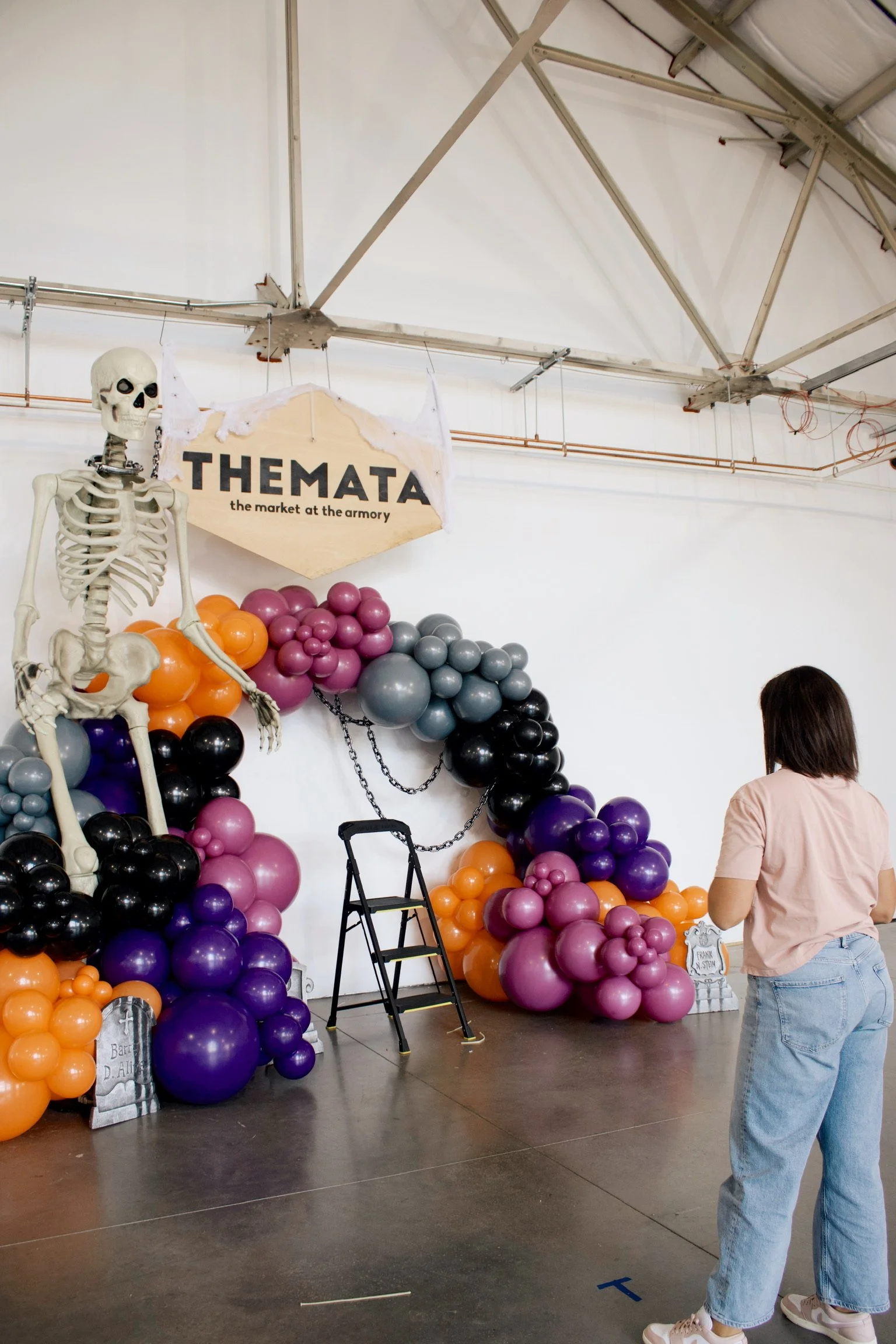 Decorations for an event featuring a skeleton figure, balloon arrangements in purple, black, orange, and pink, a sign that reads 'TH EMAT' with smaller text 'the market at the armory,' a small step ladder, and a woman with shoulder-length hair wearing a light pink shirt and jeans.