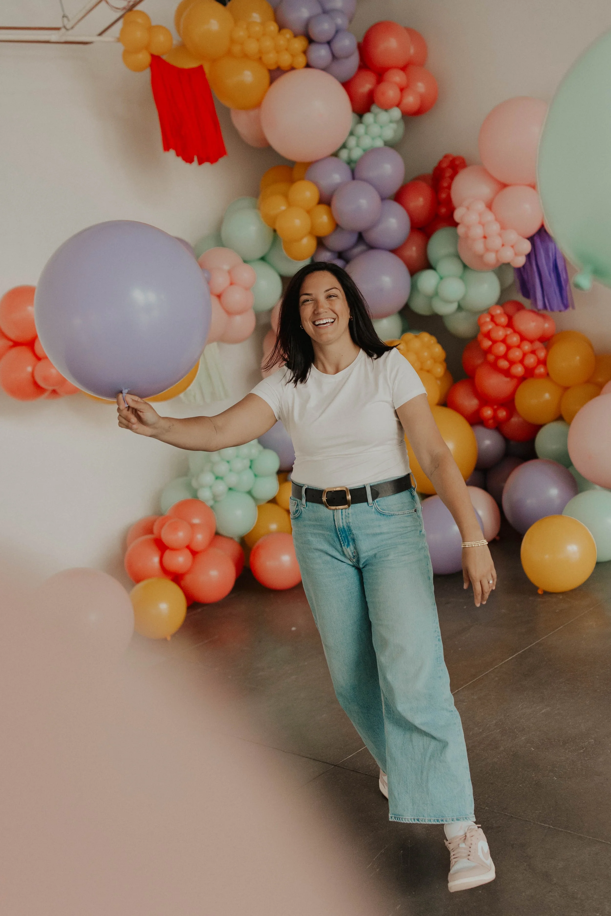 A woman smiling and holding a purple balloon with a colorful balloon decoration in the background.