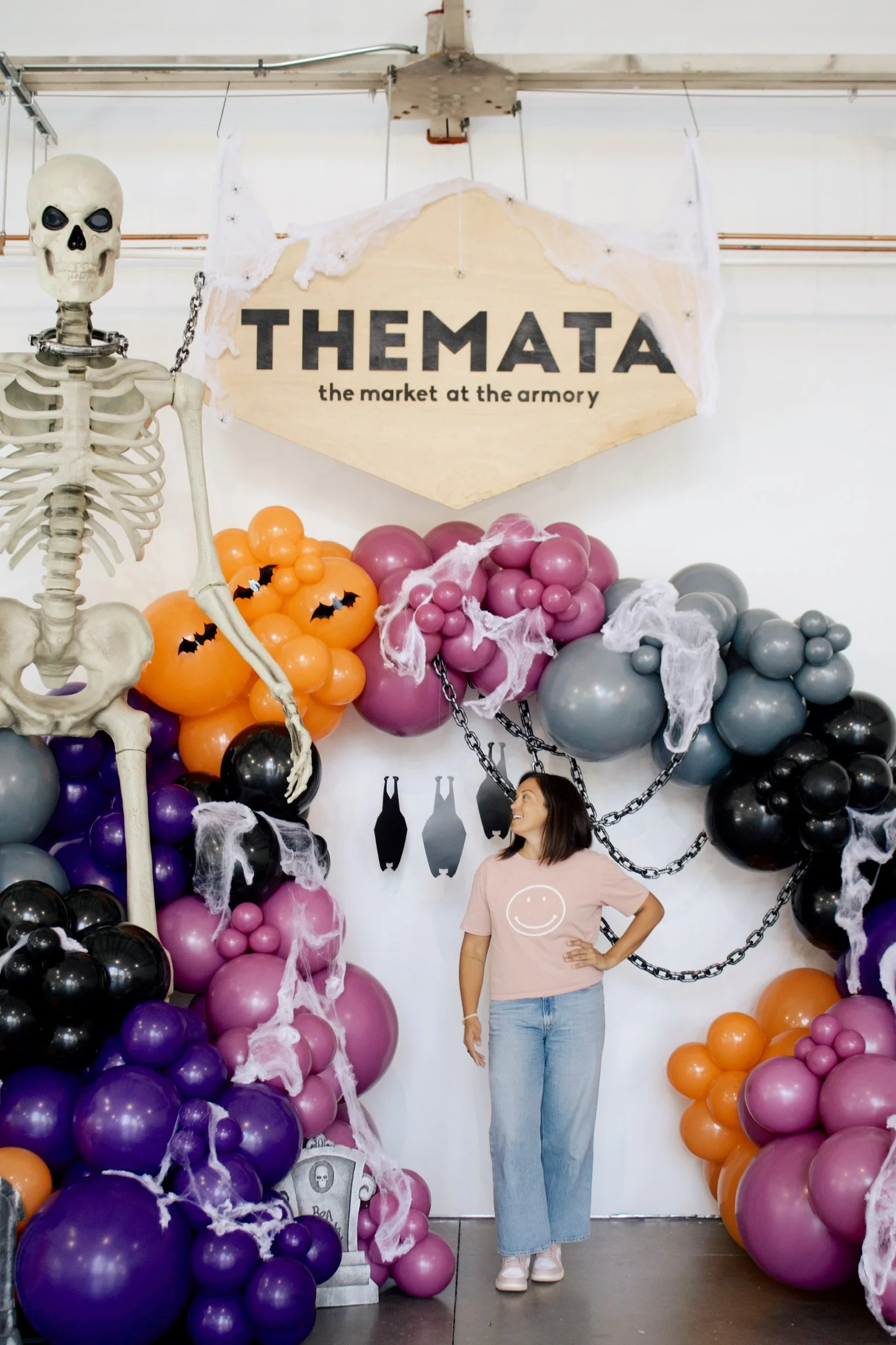 A young woman standing in front of a Halloween-themed decoration with balloons, cobwebs, chains, and a skeleton. She is wearing a light pink T-shirt with a smiley face and light blue jeans.