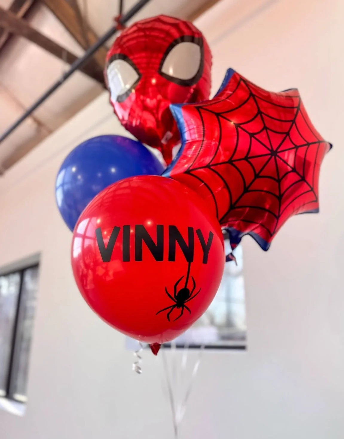 A collection of Spider-Man themed balloons, including a Spider-Man mask, a spider web design, and a red balloon with the name 'Vinny' and a spider graphic, all set against an indoor background.