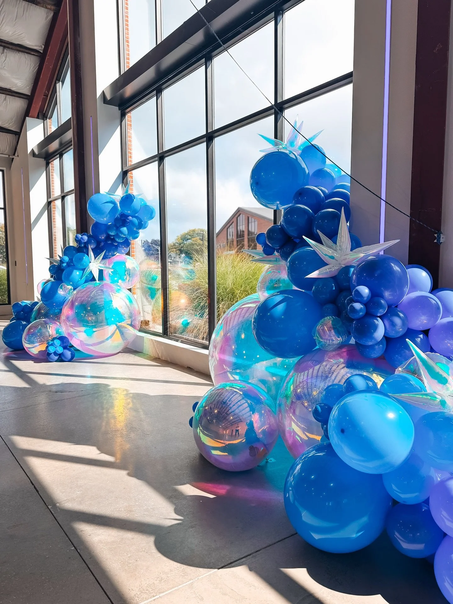Display of blue balloons and iridescent decorations inside a modern building with large windows and brick exterior visible outside.