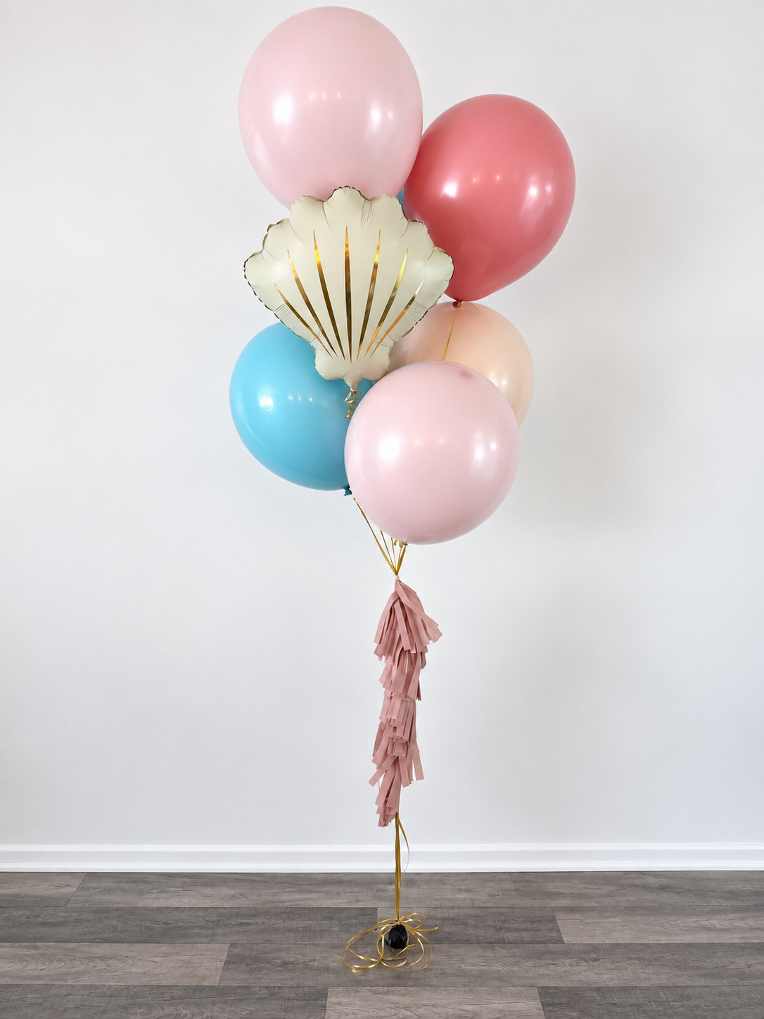 A bunch of pastel-colored balloons, including pink, blue, peach, and a gold shell-shaped balloon, tied together with pink paper fringe and gold ribbons against a plain white wall.