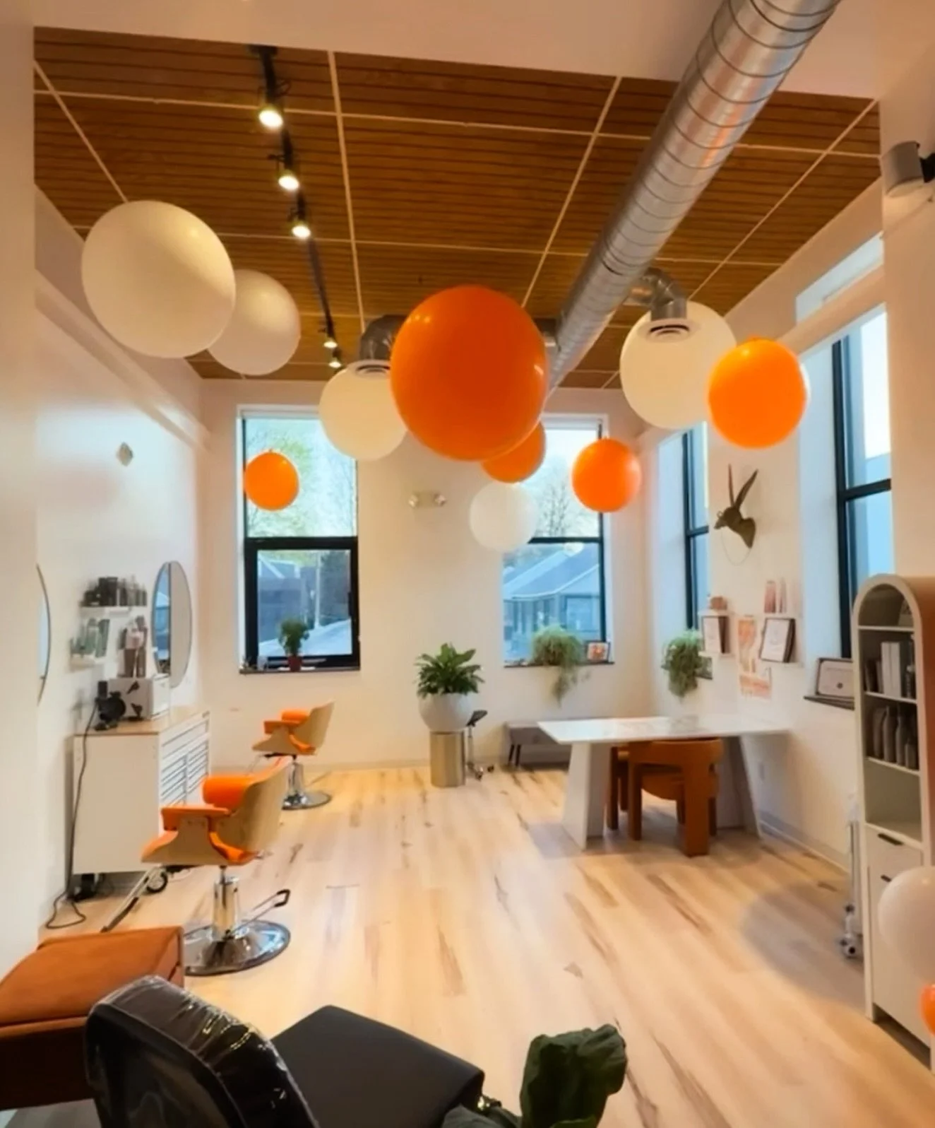 Interior of a salon with orange and white balloons hanging from the wooden ceiling, large windows, and a wooden floor. There are salon chairs, a mirror, and shelves with products.