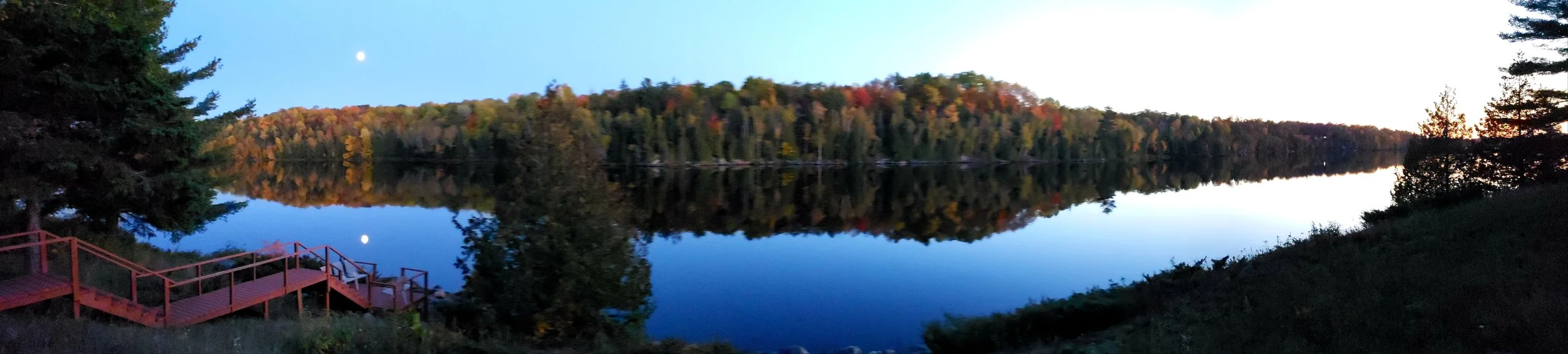 Panoramic view of a calm lake at dusk, reflecting trees with fall foliage, a wooden pier on the left, and the moon visible in the sky.