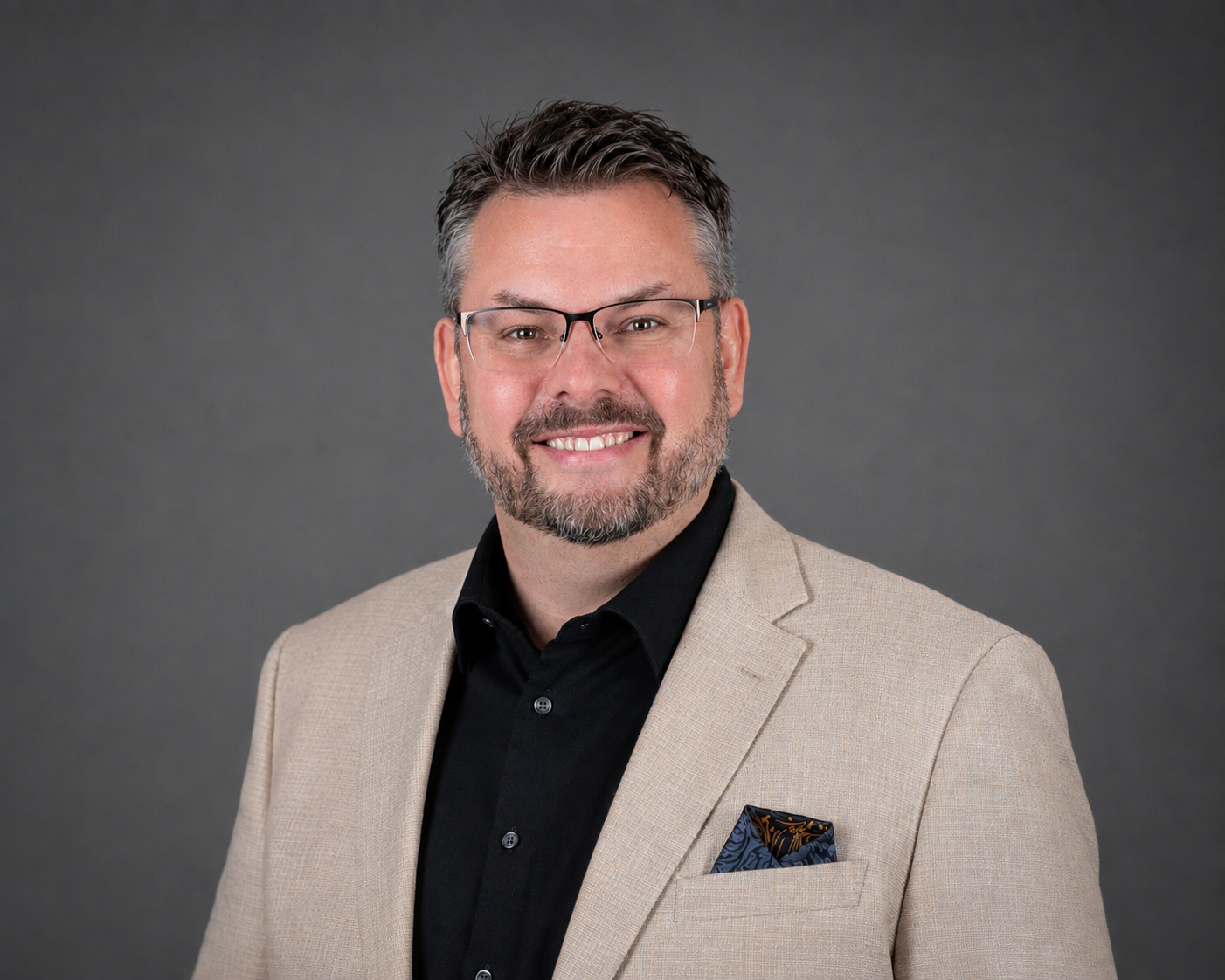 A middle-aged man with glasses, short gray hair, and a beard. He is wearing a beige blazer, black shirt, and a decorative pocket square with a dark pattern. He is smiling and standing against a dark gray background.