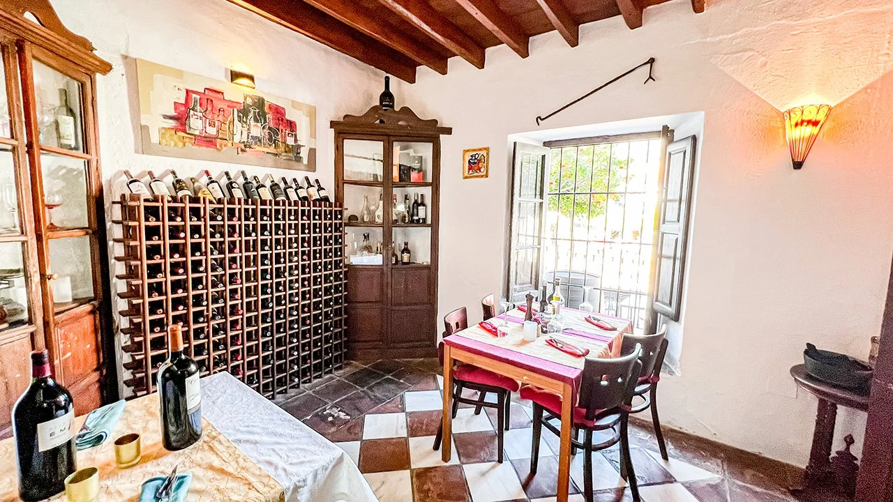 A cozy dining room with a wine rack filled with bottles, a wooden table set for four with red napkins, and a large window with open shutters letting in natural light.