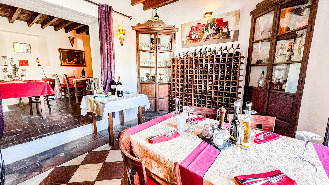 A cozy dining room with a table set for a meal, featuring a tablecloth, wine bottles, glasses, and cutlery. The room has wooden furniture, a wine rack, a cabinet with glassware, and artwork on the walls. There are purple curtains, picture frames, and