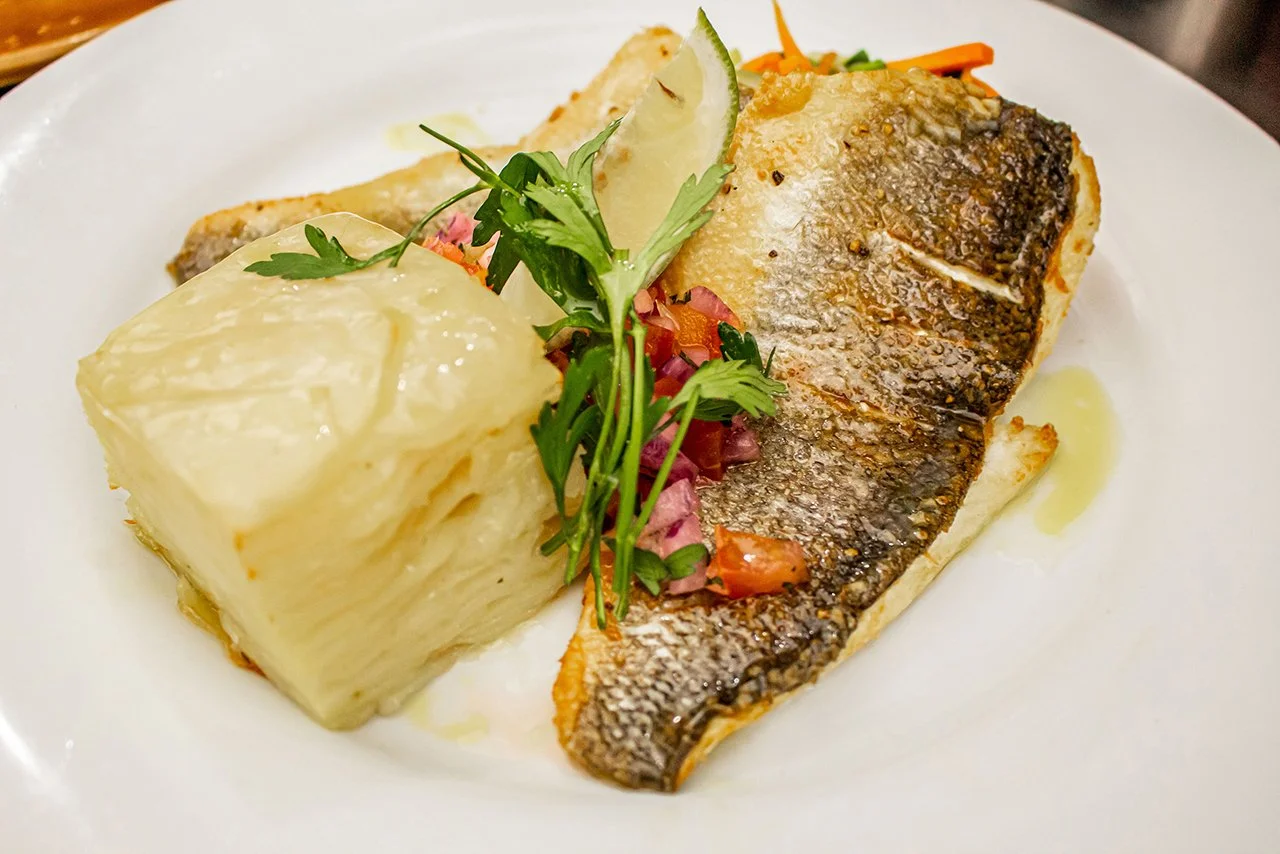 Pan-fried fish fillet with a lemon wedge, garnished with fresh herbs, served with mashed potatoes and vegetables on a white plate.
