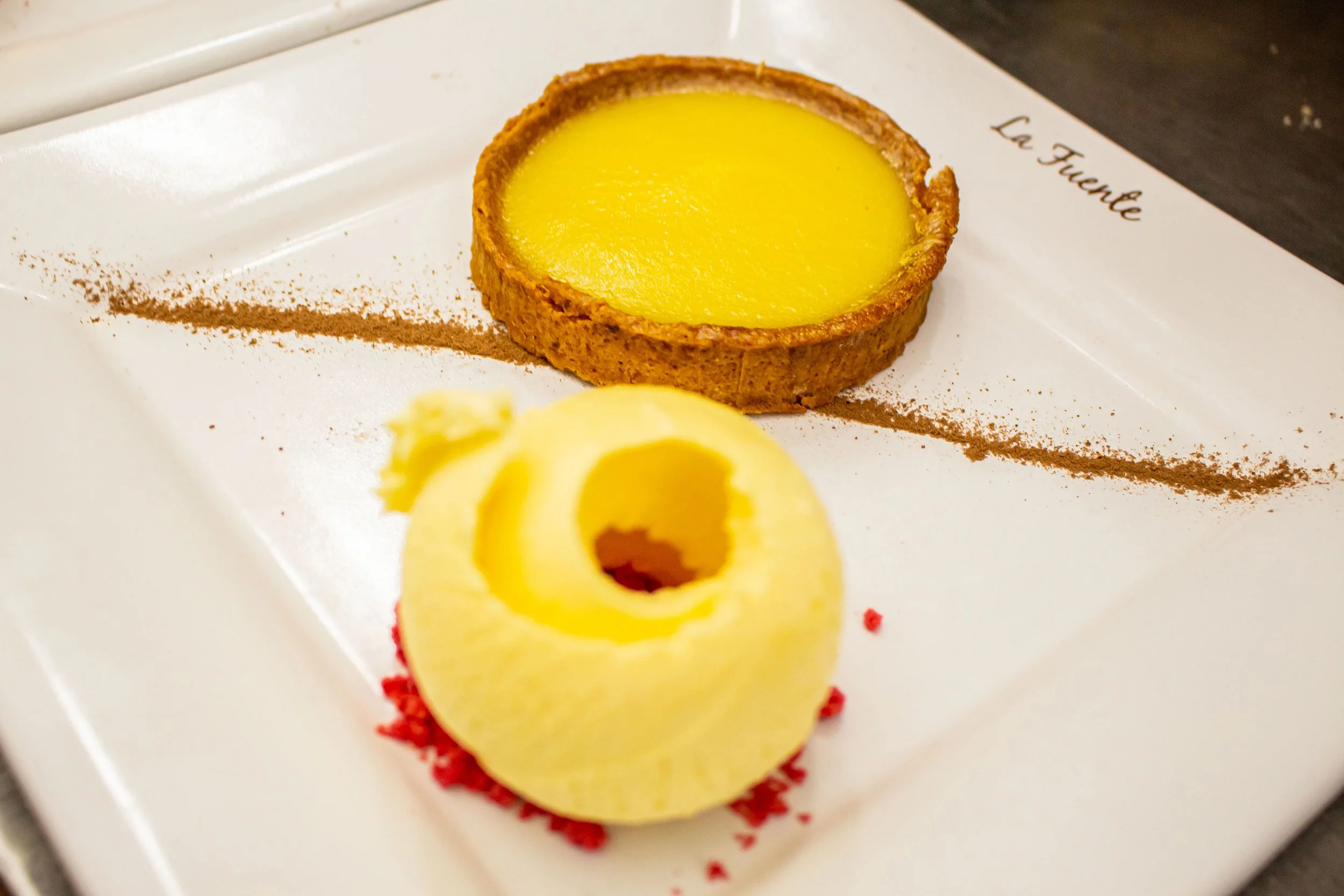 A lemon tart with a golden crust on a white plate. The tart is round and filled with bright yellow lemon custard, garnished with a lemon peel curl at the front. The plate has a thin line of cocoa powder or cinnamon dusting and the words 'Le Fvenue' w
