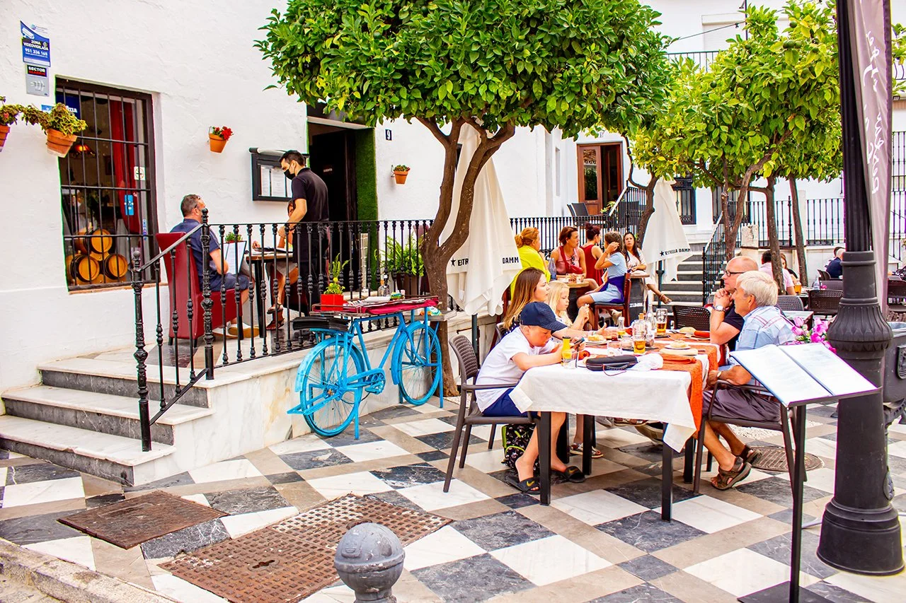 Outdoor restaurant patio with multiple tables, people dining and chatting, trees providing shade, a bright blue bicycle decoration, and a chef talking to a customer in an indoor section.