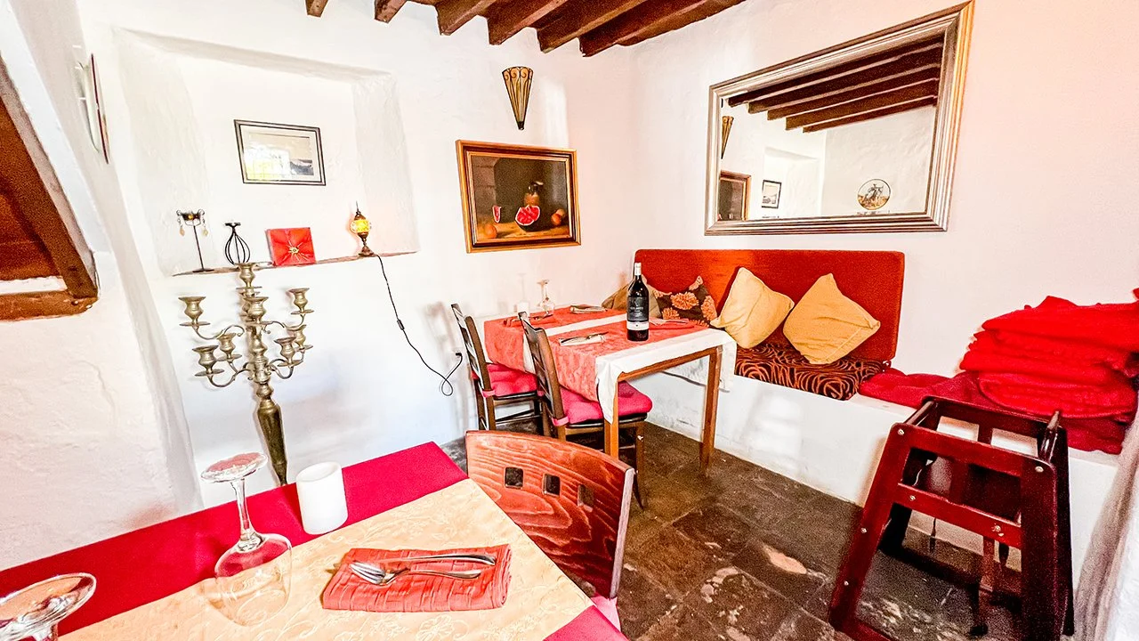 Cozy dining area with white walls, exposed wooden ceiling beams, a mirror on the wall, and a colorful table setting with a wine bottle, glasses, and utensils. There are chairs with pink cushions, a window seat with cushions, and stacked red blankets.