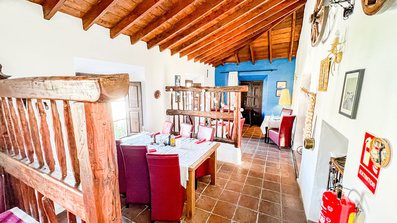 Inside a rustic restaurant or dining area with white walls, a wooden ceiling, terracotta tile flooring, and various decorations. There is a long dining table with red and white chairs, set with tableware, and a smaller table with a yellow lamp in the