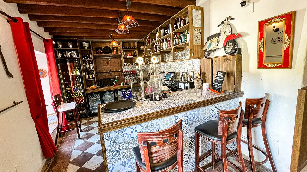 Cozy home bar with wooden and tiled details, wine bottles on shelves, vintage scooter decoration, and four wooden bar stools.