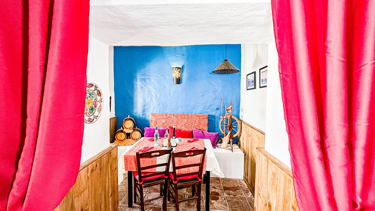 A dining room with a table, chairs, and colorful wall decor, viewed through red curtains. There is a blue accent wall and various decorative objects on the table and shelves.